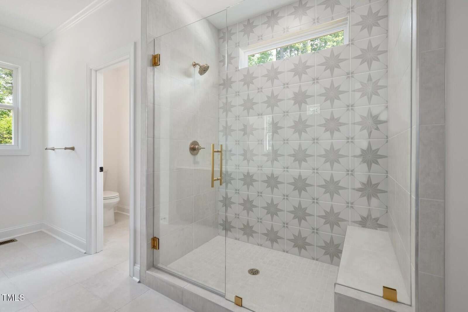 Modern bathroom with a large glass-enclosed shower featuring gold hardware and decorative star-patterned tiles. A built-in bench is inside the shower, with a window above letting in natural light. Toilet is seen in adjoining room.