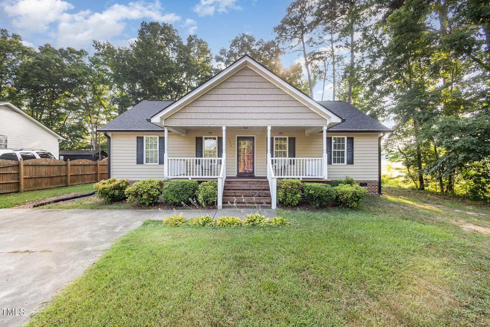 Single-story house with beige siding, a covered front porch, white railings, and steps leading to the entry door. Shrubs line the front, with a lawn and trees surrounding the property. The driveway is visible on the left.