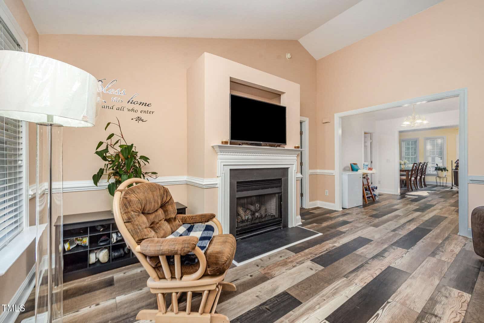 A living room with a rocking chair, fireplace with a TV above it, a potted plant, a floor lamp, and wood flooring. An open doorway leads to a dining area in the background.