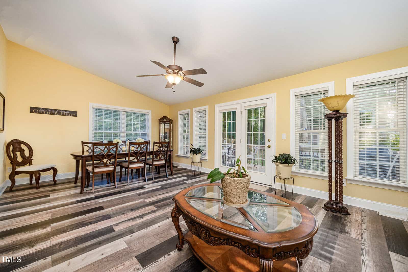 A bright dining room with yellow walls, a ceiling fan, and large windows. A wooden dining table with chairs sits near the windows, and a coffee table with a potted plant is centered on a patterned floor.