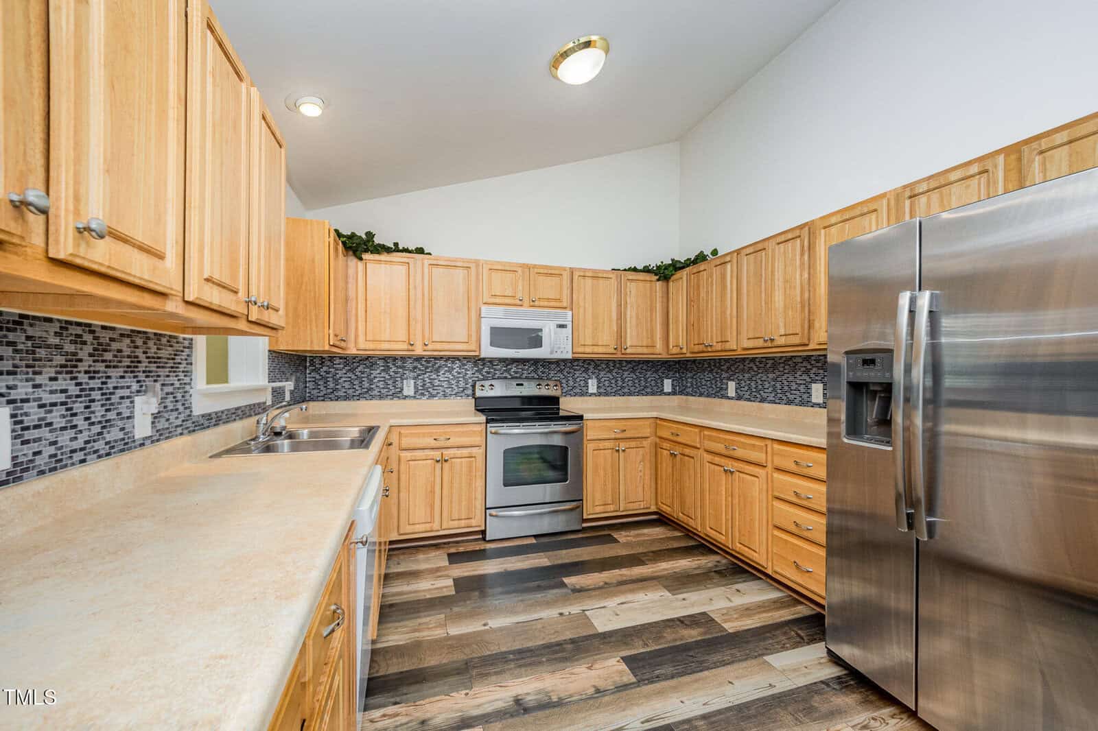 Modern kitchen with wooden cabinets, stainless steel appliances, mosaic tile backsplash, double sink, and wood-look flooring. The room has white walls, vaulted ceiling, and recessed lighting.