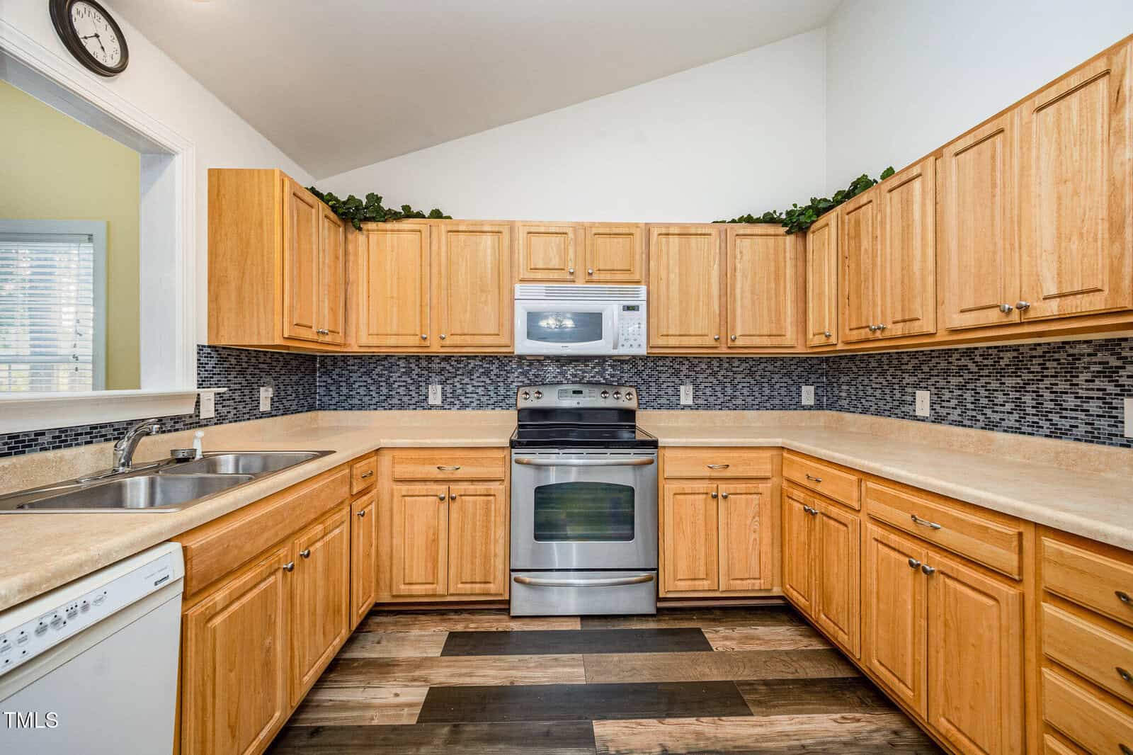 Modern kitchen with light wood cabinets, stainless steel appliances, double sink, mosaic tile backsplash, and plants above the cabinets. The floor has a mix of dark and light wood planks.