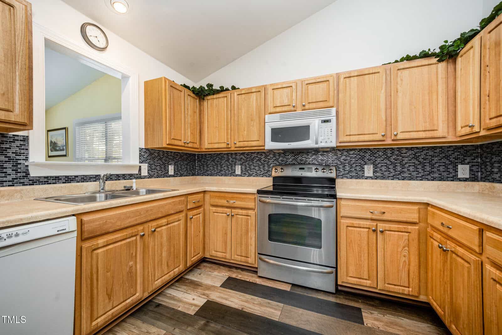 Modern kitchen with light wood cabinets, stainless steel appliances, black mosaic tile backsplash, dishwasher, double sink, beige countertops, decorative greenery above cabinets, and a clock on the wall.
