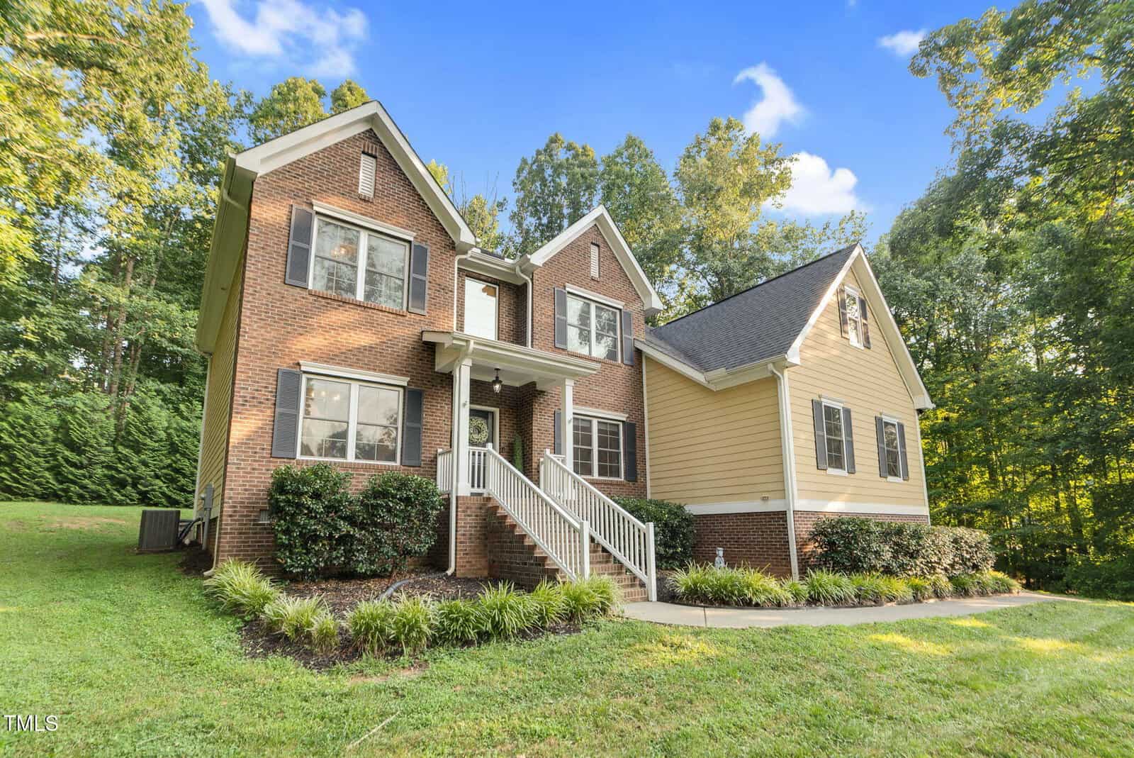 A two-story house at 3650 Pleasants Ridge Drive with red brick and cream siding, black shutters, and a gable roof, surrounded by green grass, shrubs, and trees under a blue Wake Forest sky with fluffy clouds.