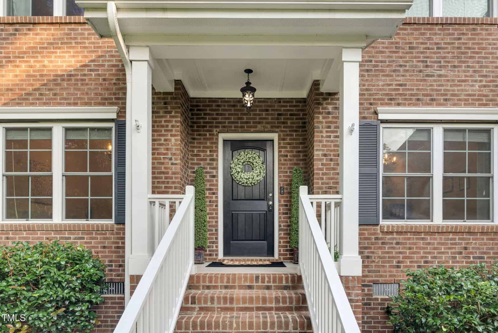 Front porch of a brick house with a black door decorated with a wreath, flanked by two narrow windows and topiary plants. White railings line the steps leading to the entrance.