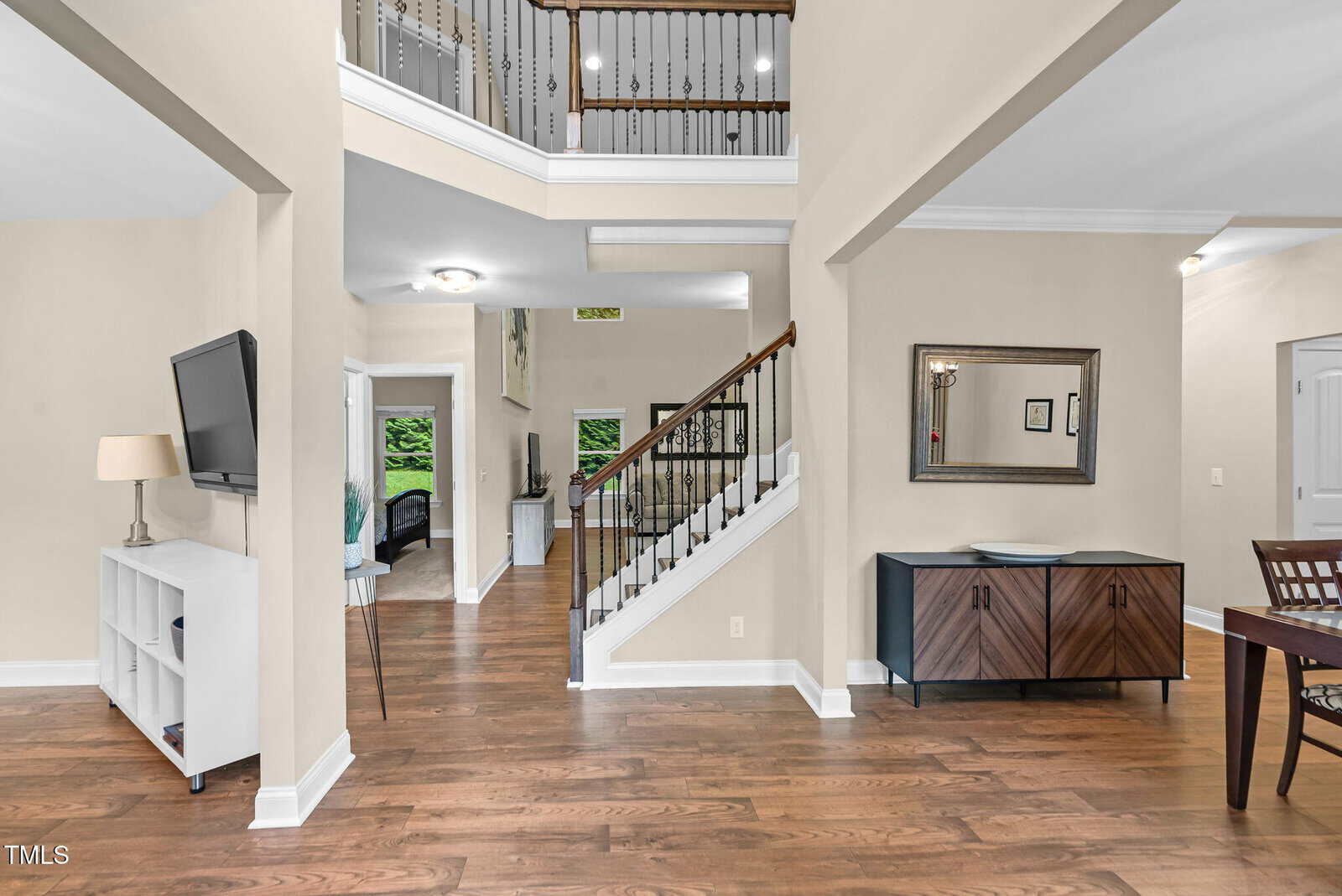 Modern home interior with hardwood floors, neutral walls, a staircase with a dark railing, wall-mounted TV, white shelves, a console table with a mirror, and a glimpse into a dining area and another room.