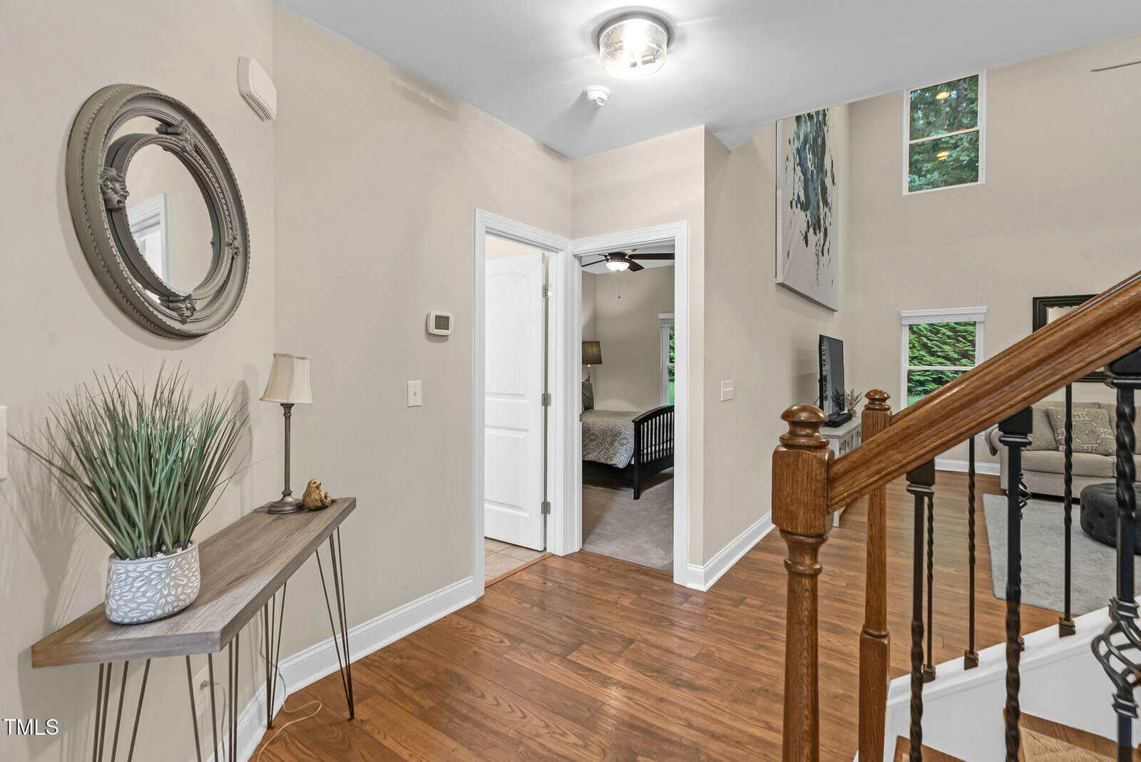 A hallway with wood floors, a console table with decor, a round wall mirror, stairs with a wood handrail, and a view into a bedroom and a living area with large windows.