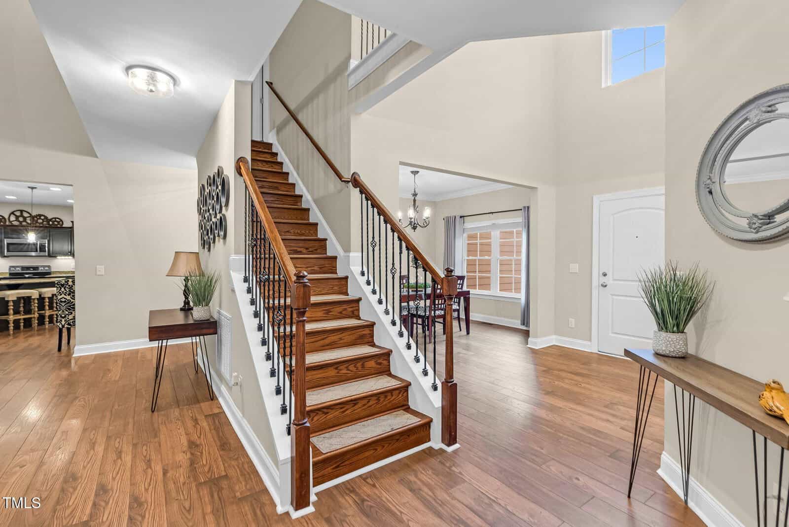 Bright entryway with wooden staircase, metal railings, hardwood floors, and modern decor. Dining area and kitchen are visible, with a round mirror on the wall and plants on tables. Large window provides natural light.