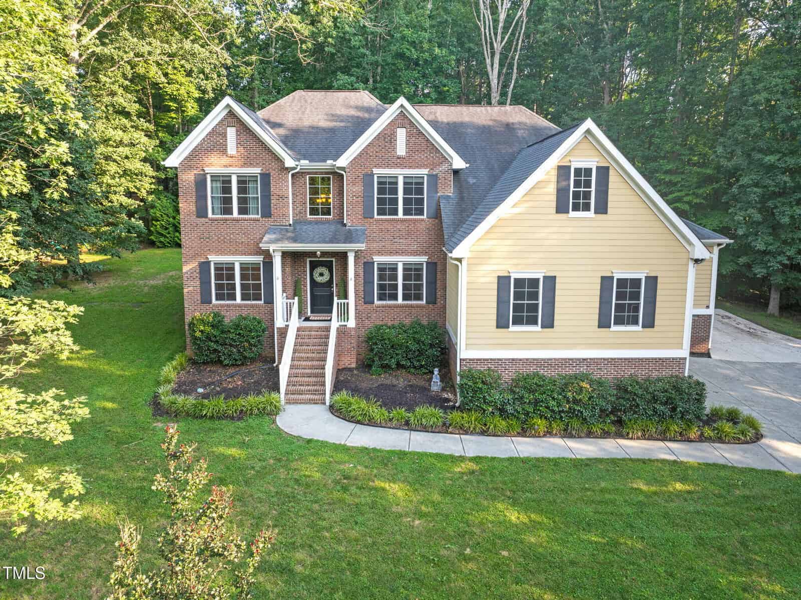 A two-story house with red brick and yellow siding, black shutters, a gable roof, front steps leading to a covered entry, landscaped bushes, and a curved driveway, surrounded by green trees and grass.