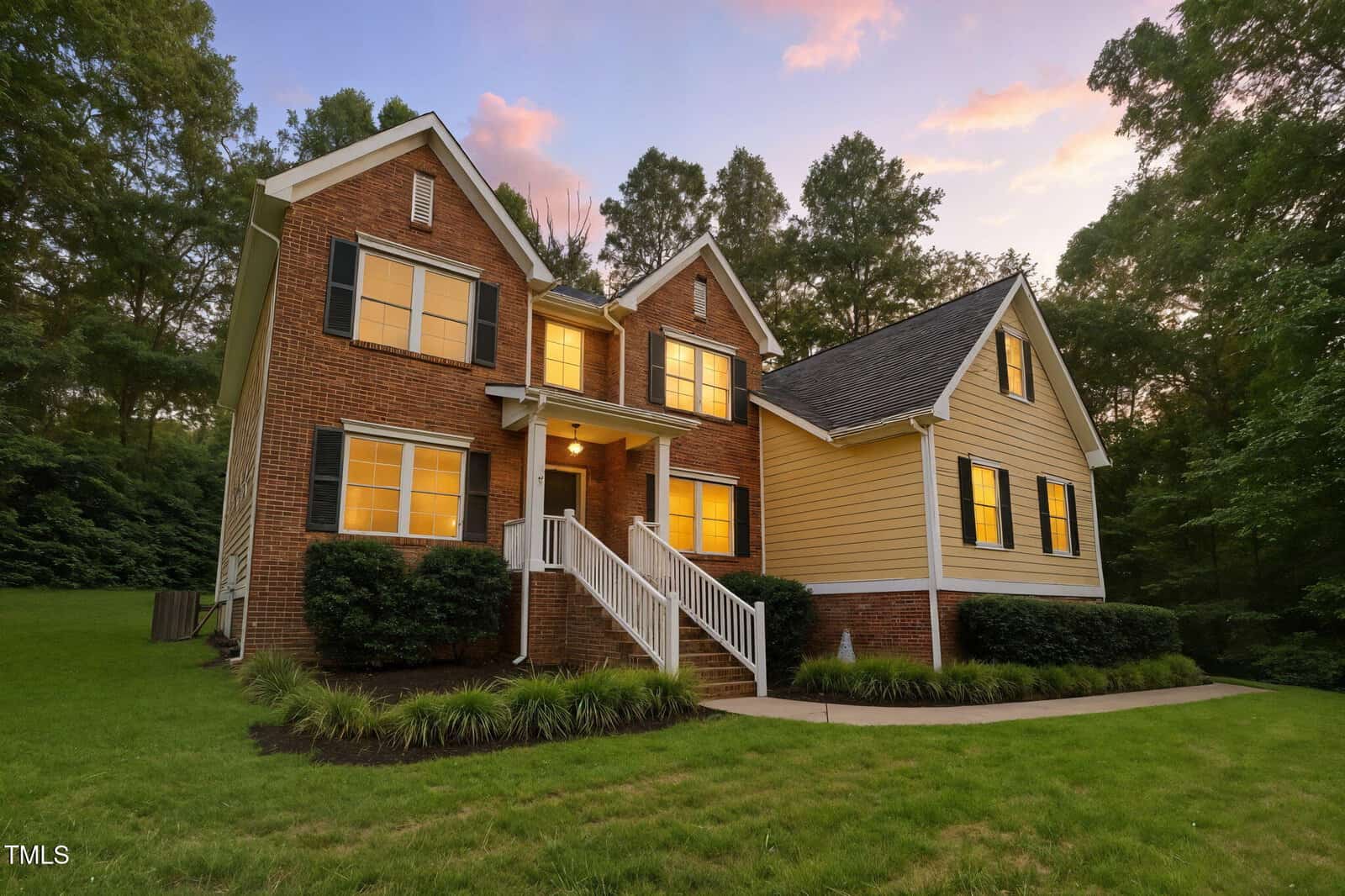 A two-story house with brick and yellow siding, black shutters, and a front porch with white railings, set on a green lawn with trees in the background at sunset.