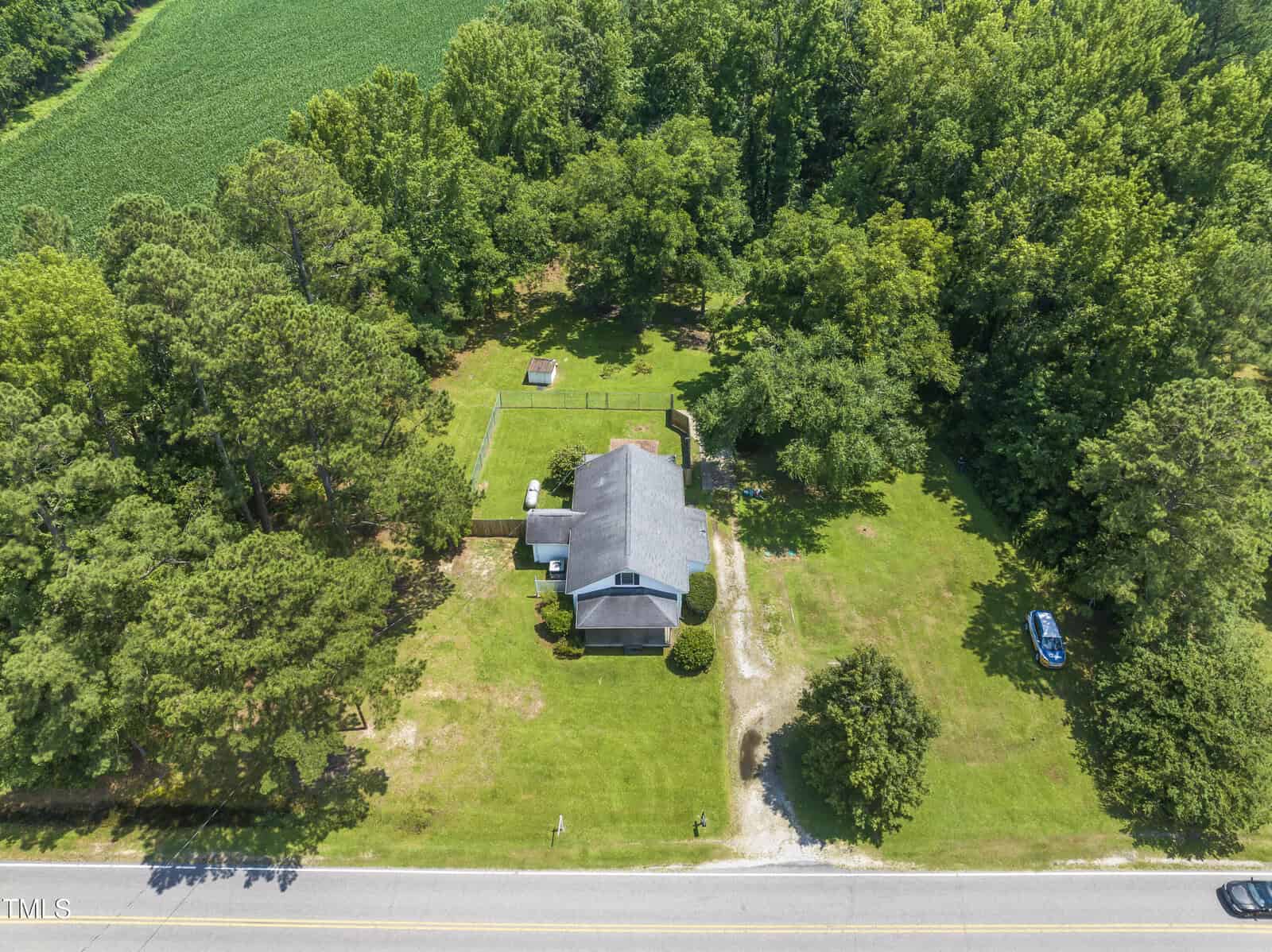 Aerial view of a house surrounded by trees, a grassy yard, and a driveway leading to a road. There is a car parked on the right side near the edge of the property. A field is visible in the upper left corner.