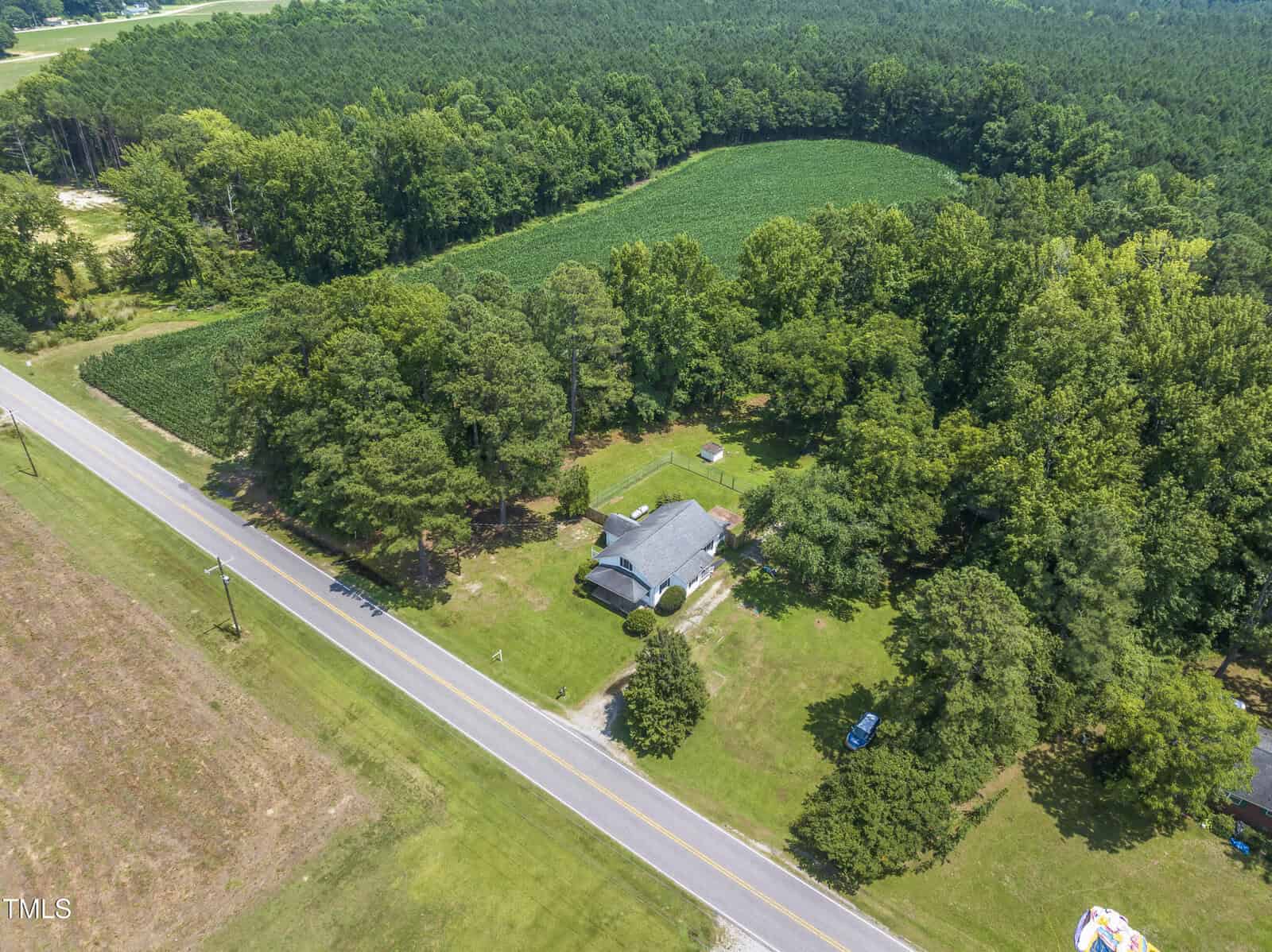 Aerial view of a house surrounded by trees and open fields, located next to a rural road with dense forest and farmland in the background.