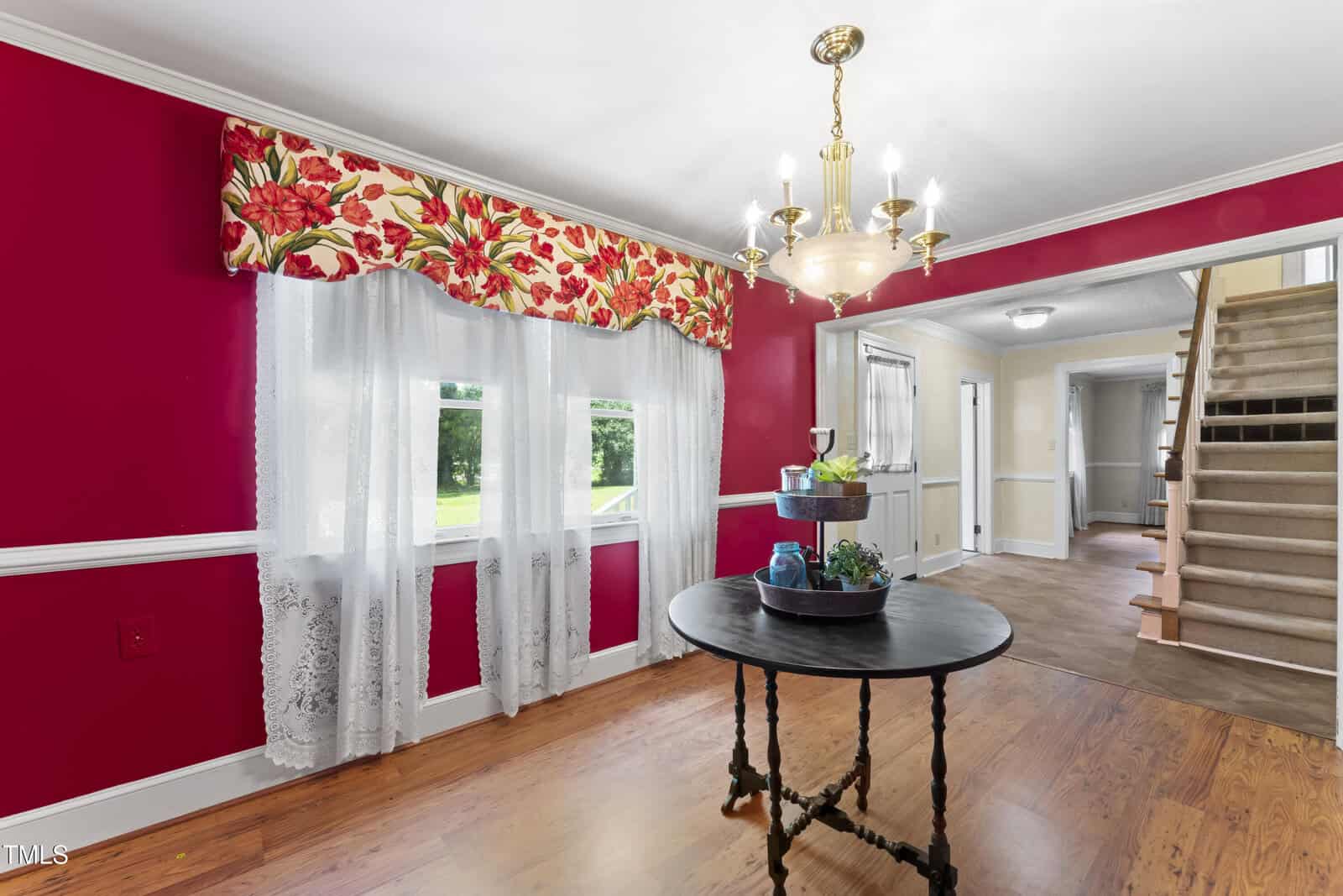 A dining room with bright red walls, lace curtains with a floral valance, a round black table with decorative items, a brass chandelier, wood flooring, and a staircase leading to the upper floor.