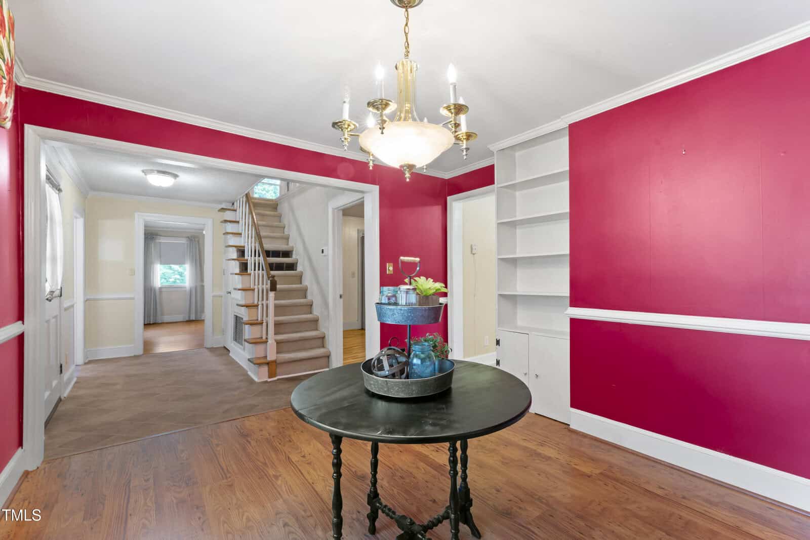 A room with red walls, white trim, built-in shelves, wood flooring, and a round black table under a brass chandelier. A staircase and two doorways are visible in the background.