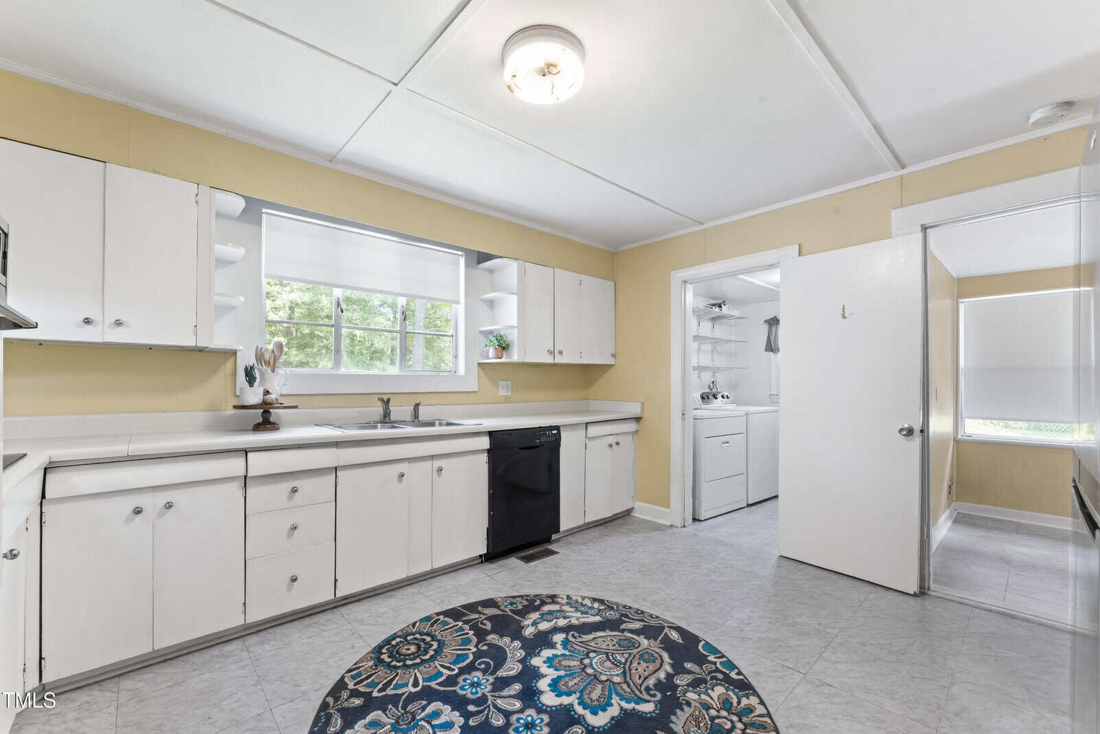 A kitchen with white cabinets, a black dishwasher, and a window above the sink. A round patterned rug is on the floor. The kitchen opens to a laundry area and has yellow walls with white trim.