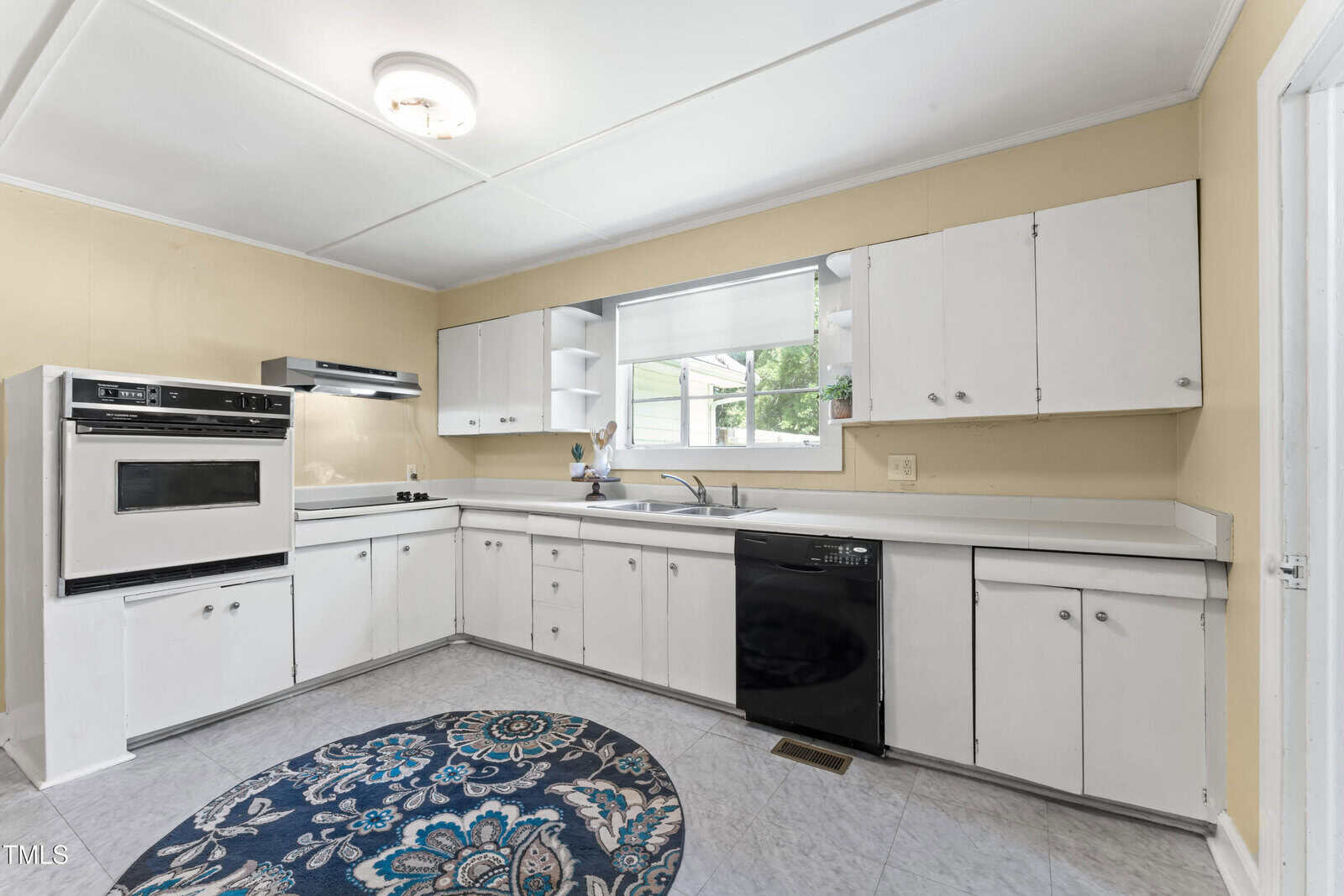 A kitchen with white cabinets, a black dishwasher, a built-in oven, a cooktop, and a double sink under a window. There is a blue patterned rug on the light tile floor.