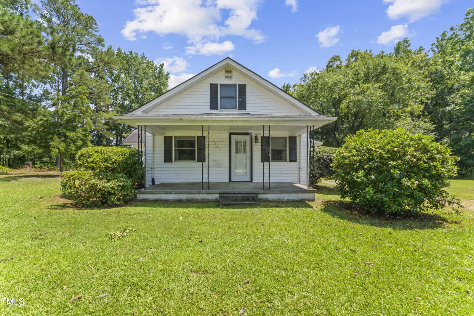 A small white house with a front porch, steps leading up, and two bushes on either side. The house sits on a green lawn with trees in the background under a blue sky with scattered clouds.