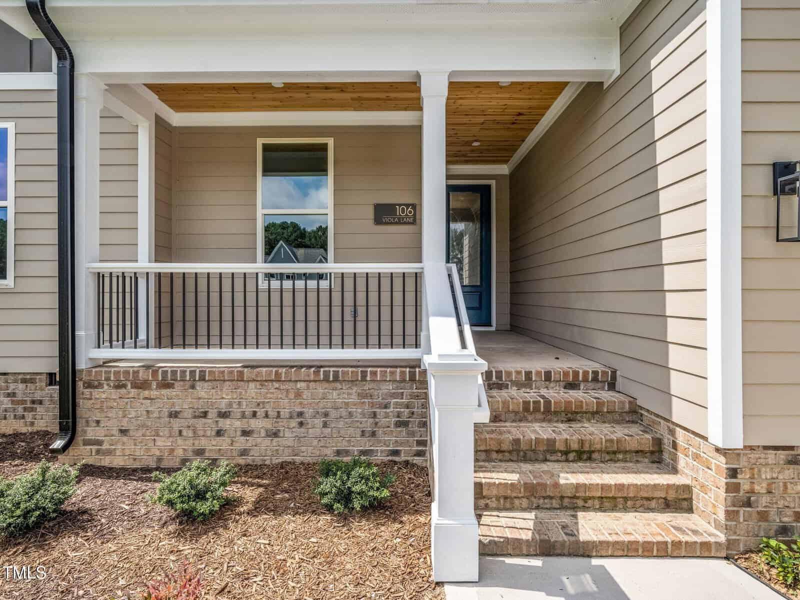 Covered front porch of a house with beige horizontal siding, brick steps, black metal railing, and a wood-paneled ceiling. There is a window, a door with glass, and a small landscaped area with shrubs.