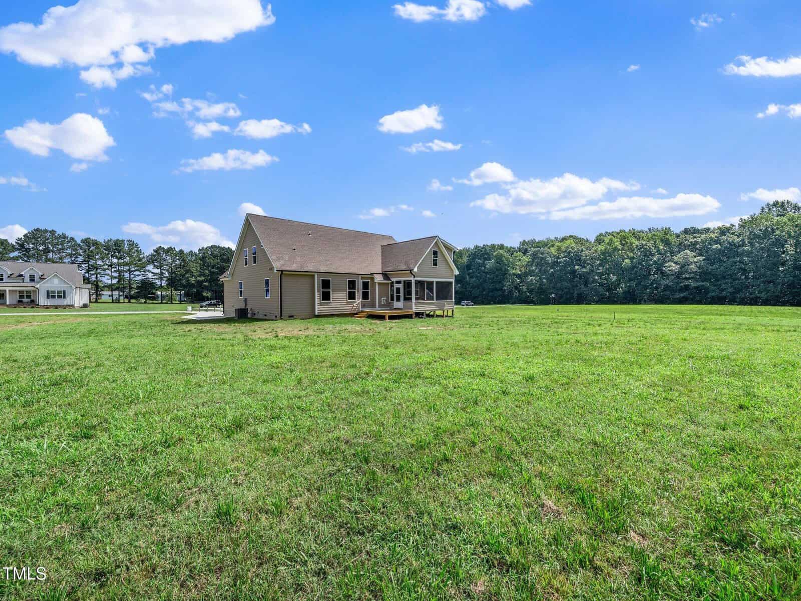 A tan house with a sloped roof sits on a large, grassy lawn under a blue sky with scattered clouds. Trees line the background, and another house is visible in the distance to the left.