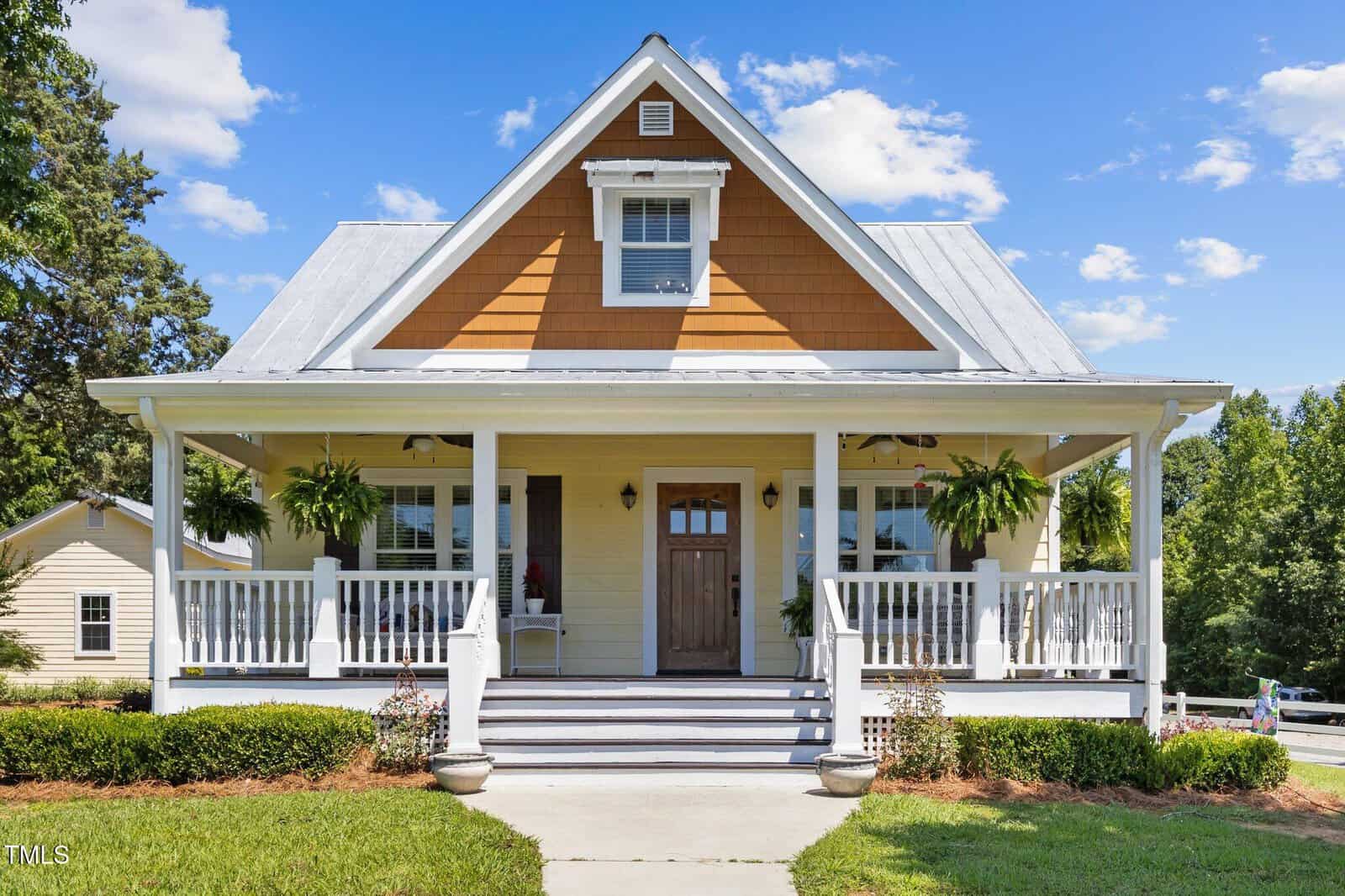 A yellow house with white trim at 4050 Culbreth Road has a wooden front door, two large windows, a gabled roof, covered porch with hanging plants, and steps to a concrete walkway. Greenery and shrubs line the front yard in scenic Stem.