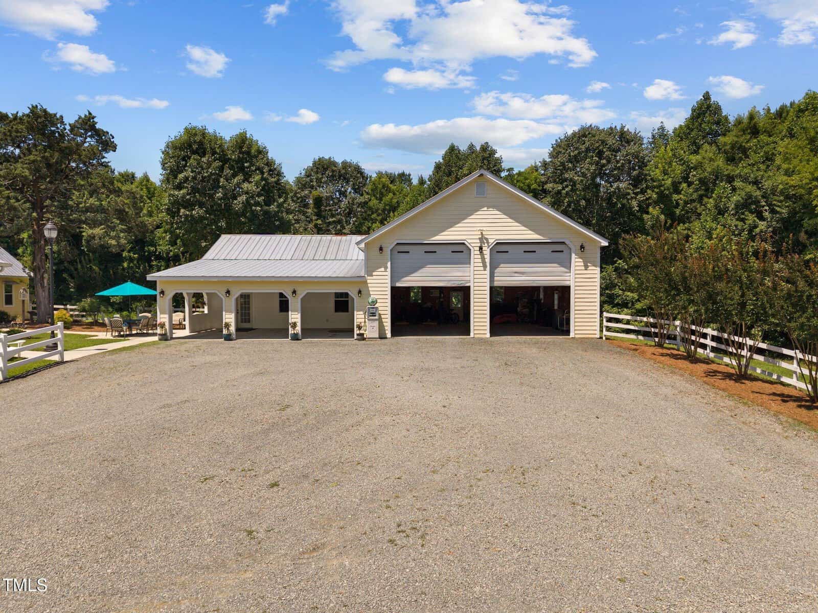 A large beige building with two garage doors and a covered porch sits on a gravel driveway, surrounded by white fencing, trees, and greenery under a partly cloudy sky.