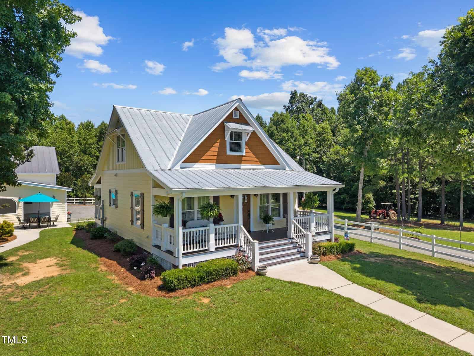 A yellow two-story house with a metal roof and front porch sits on a well-kept lawn. The house is surrounded by trees, a white fence, and a curved sidewalk leading to the entrance.