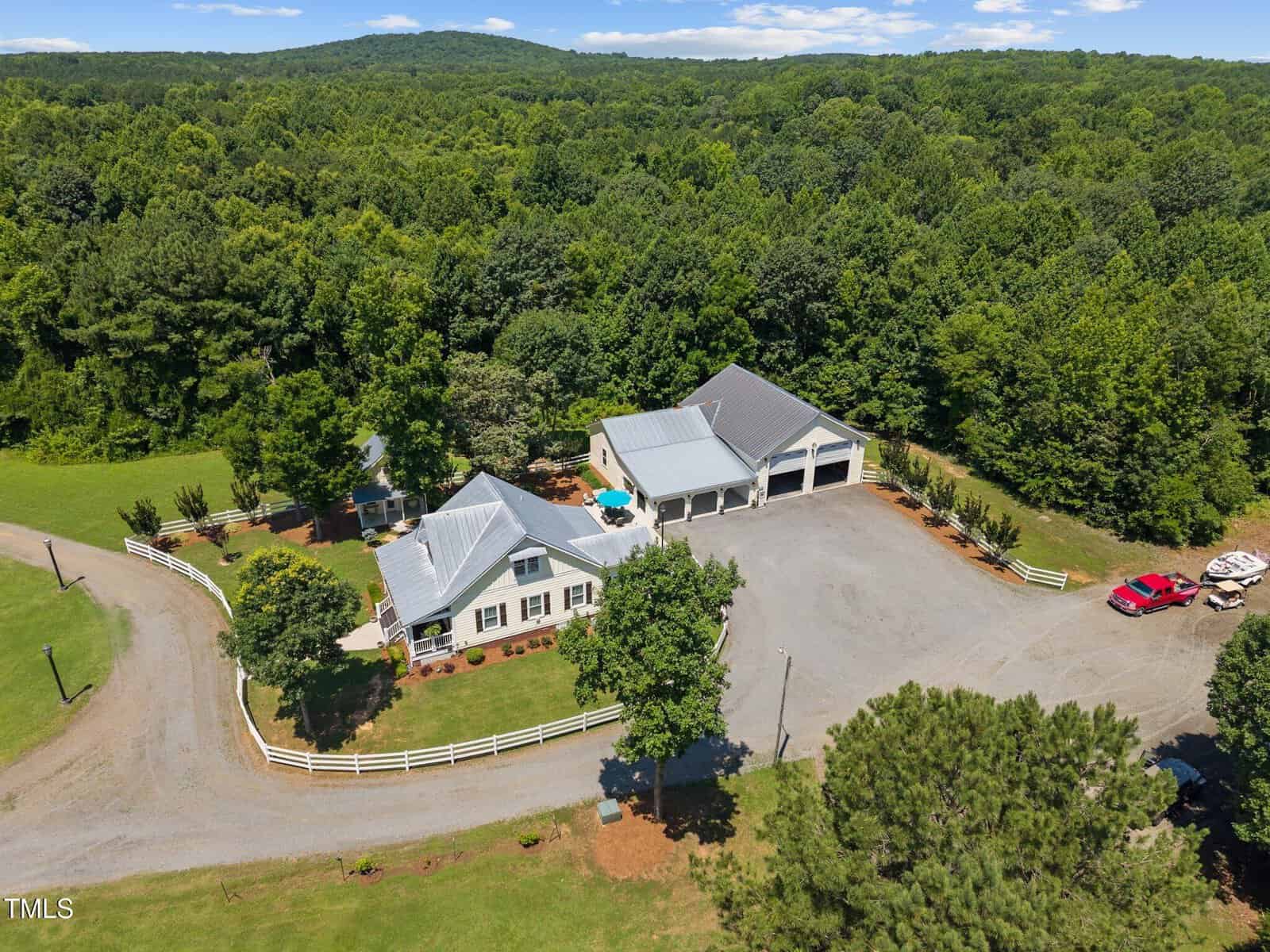 Aerial view of a white country house with a metal roof, a circular driveway, a detached garage, and a fenced yard surrounded by dense trees. Several vehicles are parked near the buildings.