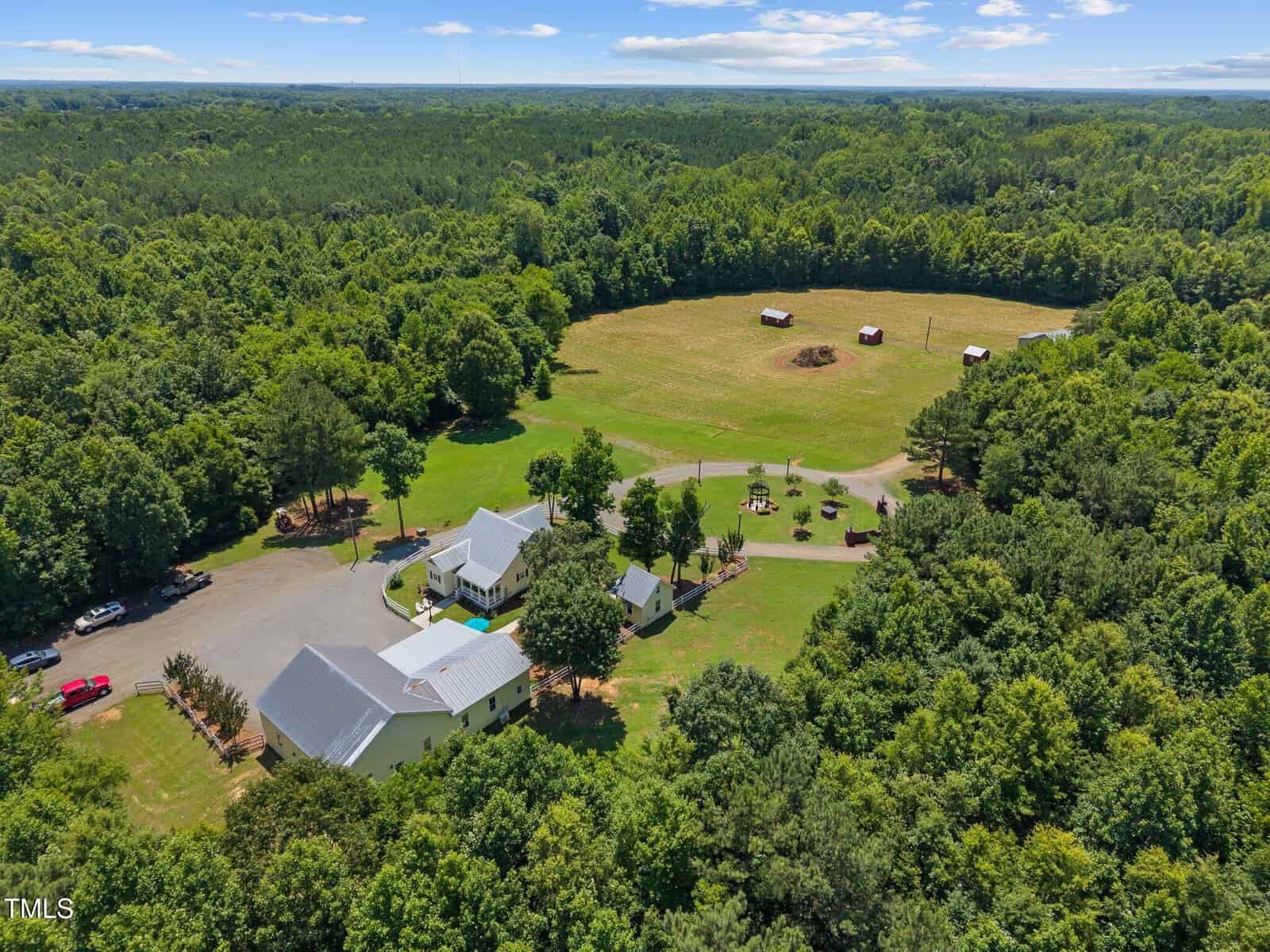 Aerial view of a rural property with multiple white buildings, a gravel driveway, several vehicles, open grassy fields, and a few small barns or sheds, surrounded by dense green forest under a partly cloudy sky.