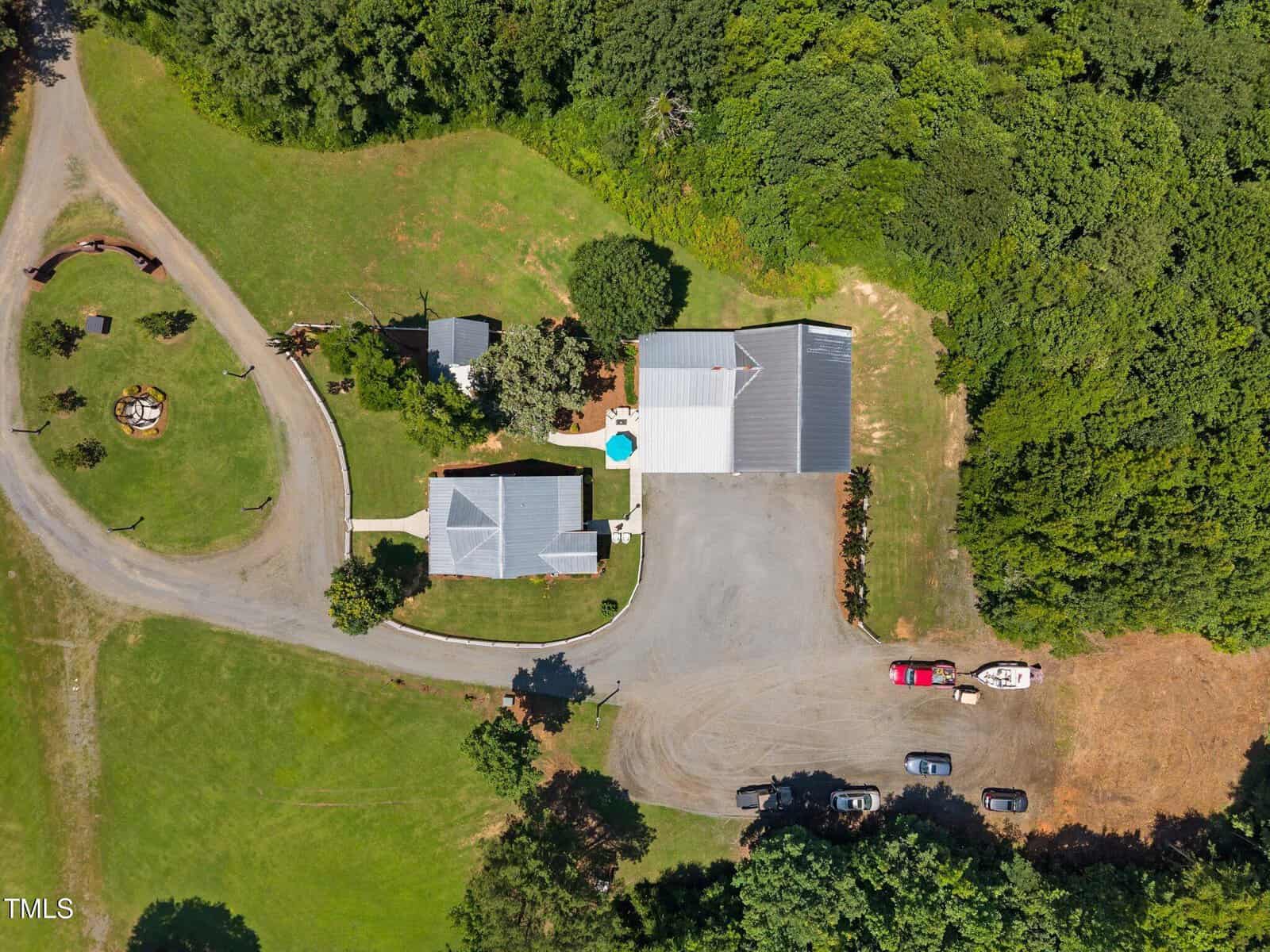 Aerial view of a property with two main buildings, a circular driveway, a small pool, several parked vehicles, and large grassy areas surrounded by trees and forest.