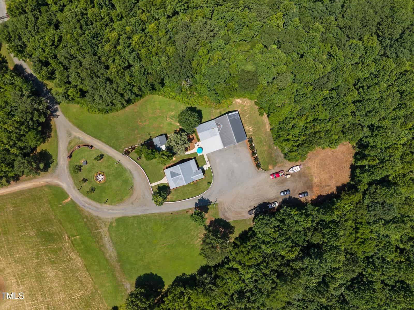 Aerial view of a rural property with two buildings, a driveway, several parked vehicles, a circular seating area with a fire pit, and surrounding trees and grassy areas.