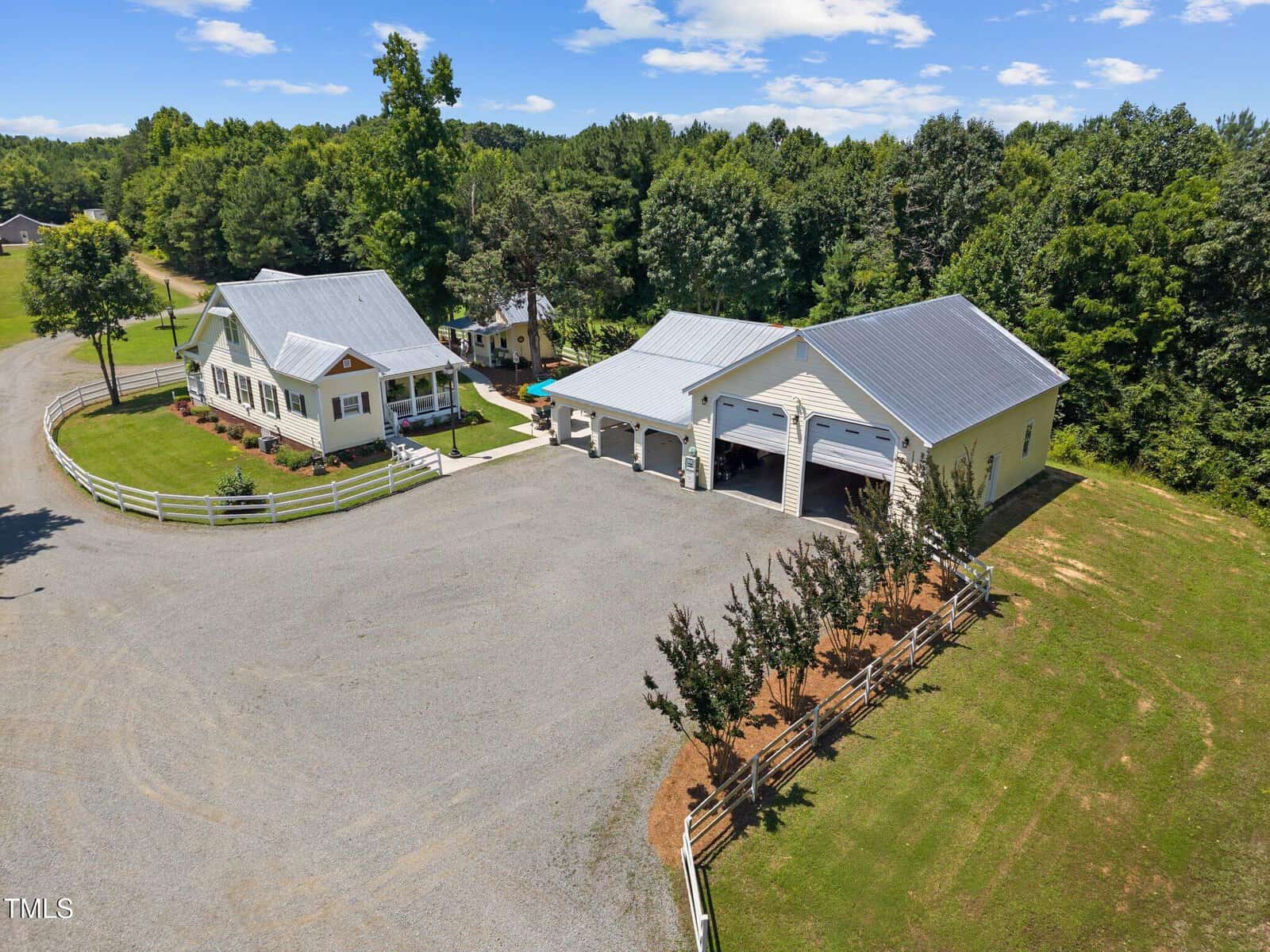 Aerial view of a rural property with a white single-story house, a large detached garage with three doors, a gravel driveway, a fenced yard, and surrounding trees under a clear blue sky.