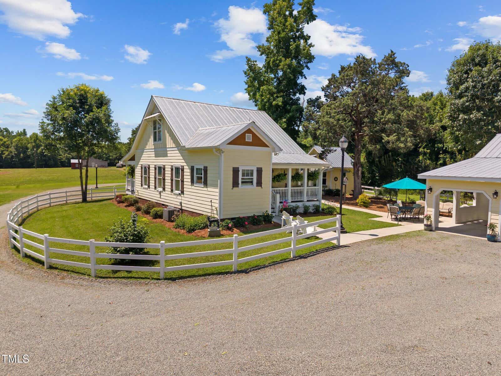 A yellow house with a metal roof and white trim sits in a landscaped yard with a white fence, surrounded by trees and open grass under a blue sky with scattered clouds. A covered patio is visible on the right.