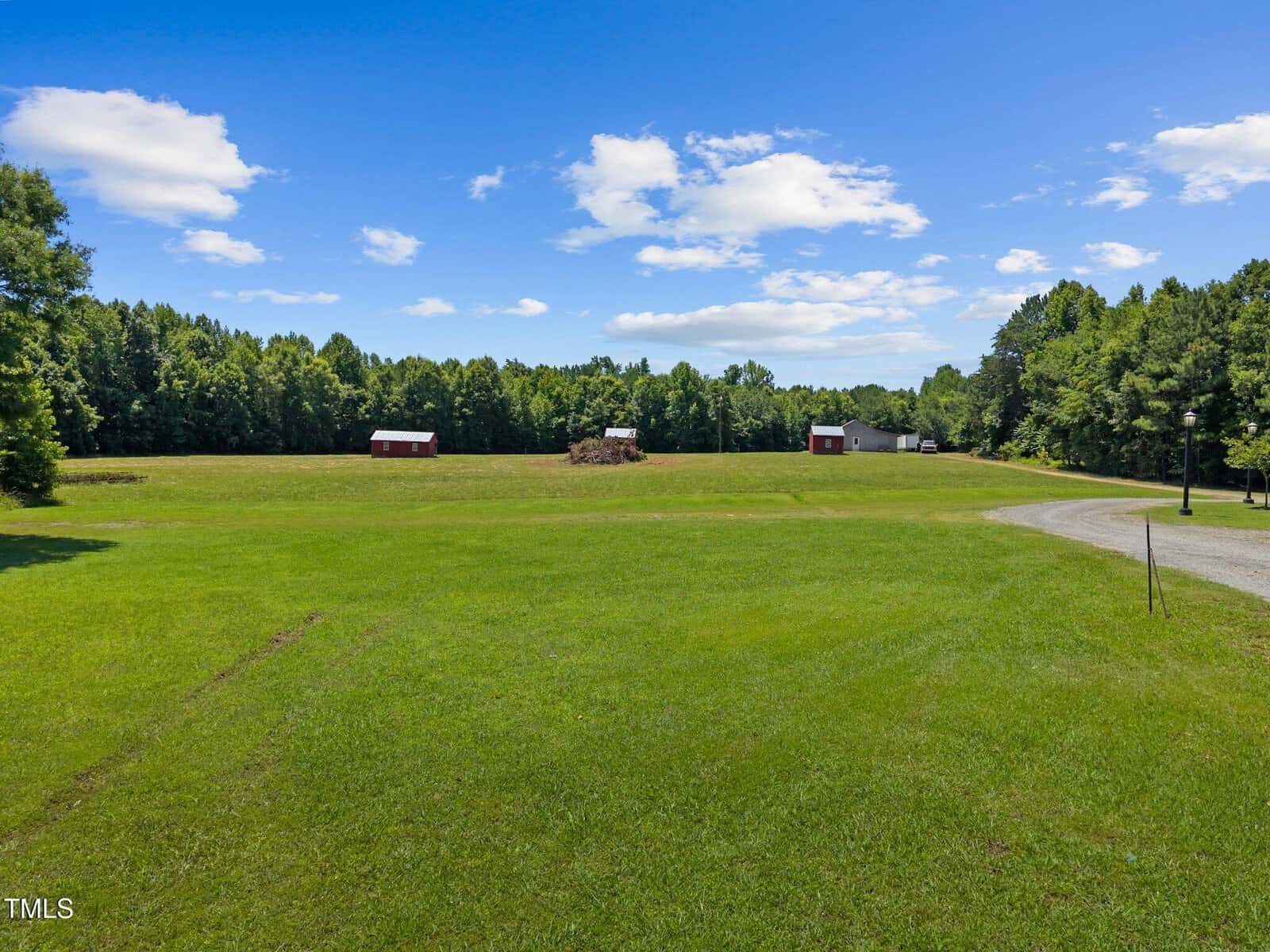 A large, open grassy field with three small red and white barns spread across the landscape; bordered by trees under a blue sky with scattered clouds and a gravel driveway curving through the right side.