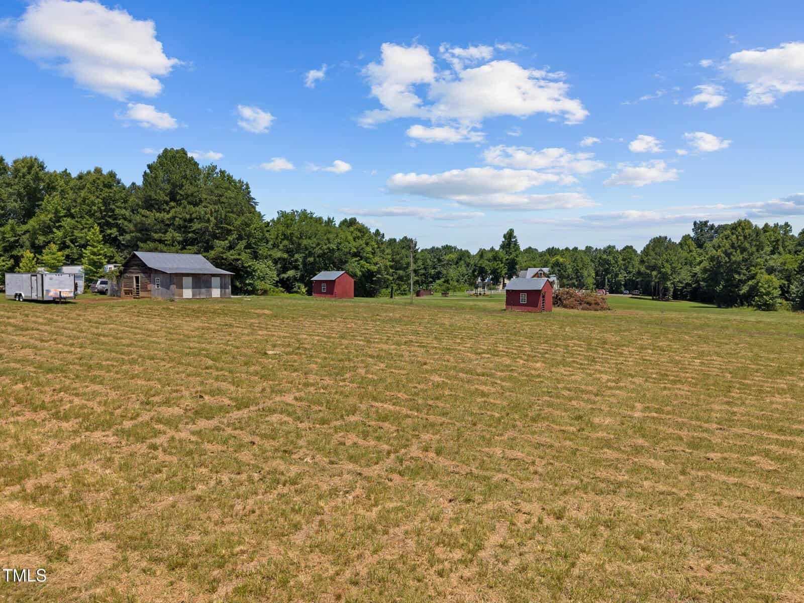 A large grassy field with scattered small red and gray sheds, surrounded by dense green trees under a blue sky with scattered clouds.