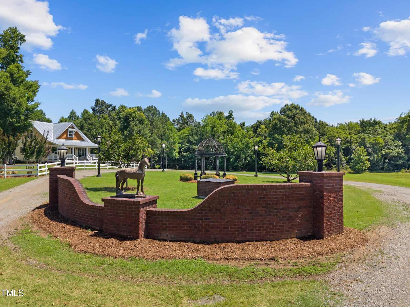 A curved red brick wall with two lanterns and a horse statue stands at a gravel driveway entrance. Behind it is green grass, trees, a gazebo, and a white farmhouse under a blue sky with fluffy clouds.
