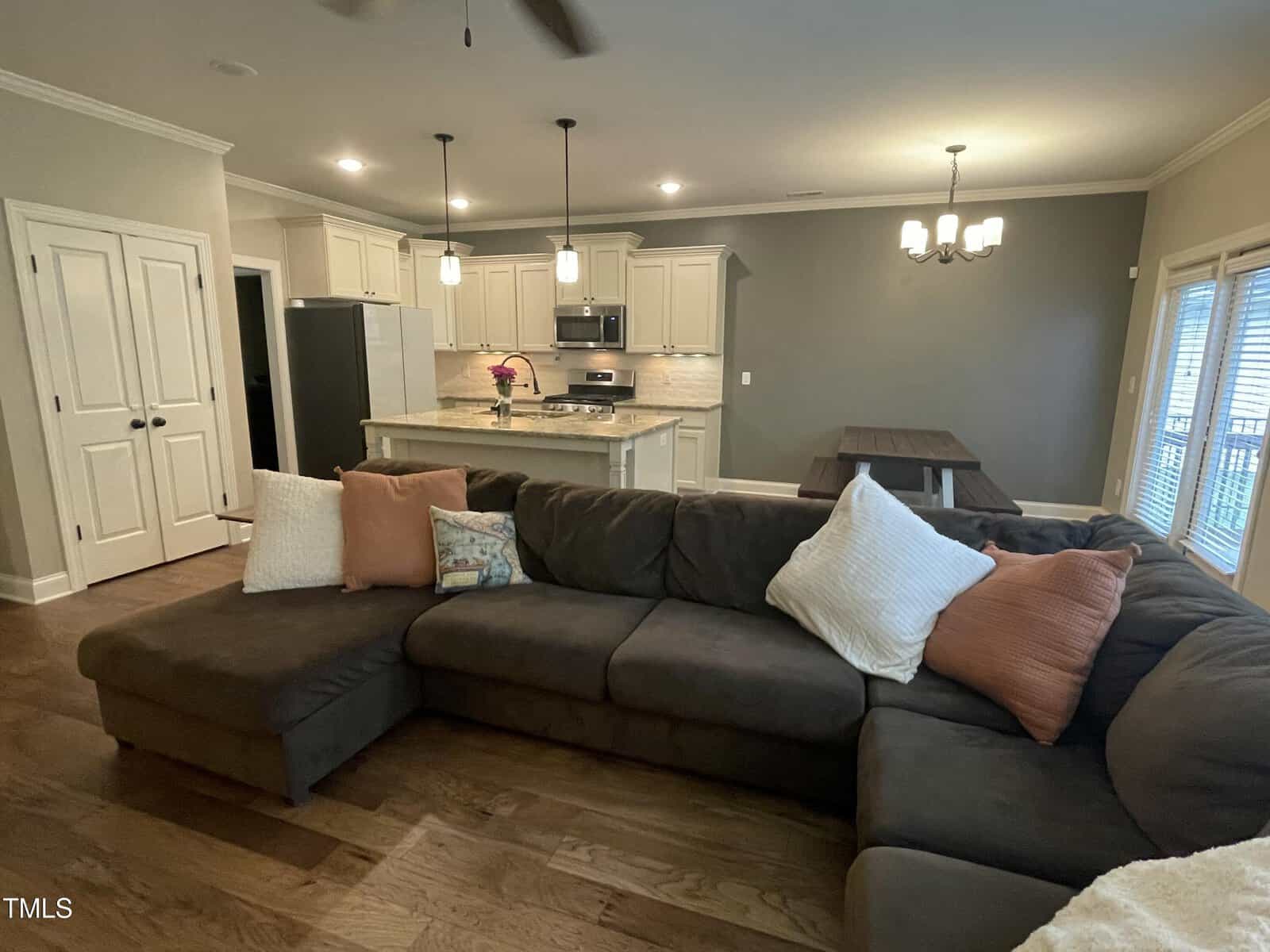 A modern kitchen with white cabinets and stainless steel appliances is shown behind a brown sectional sofa with white and orange pillows. A wooden dining table sits near large windows on the right. The room features hardwood floors.