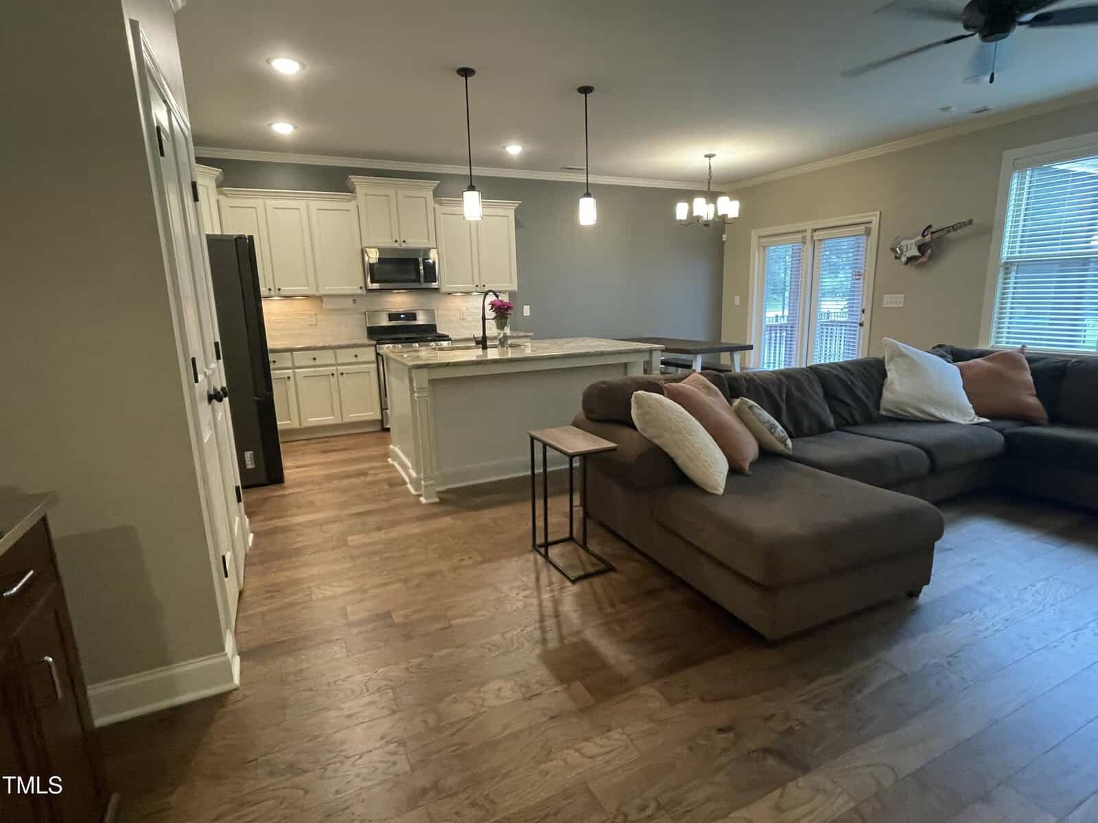 Open-concept living area with a brown sectional sofa, cushions, and side tables in front of a kitchen with white cabinets, stainless steel appliances, a large island, and pendant lights. Hardwood floors throughout.