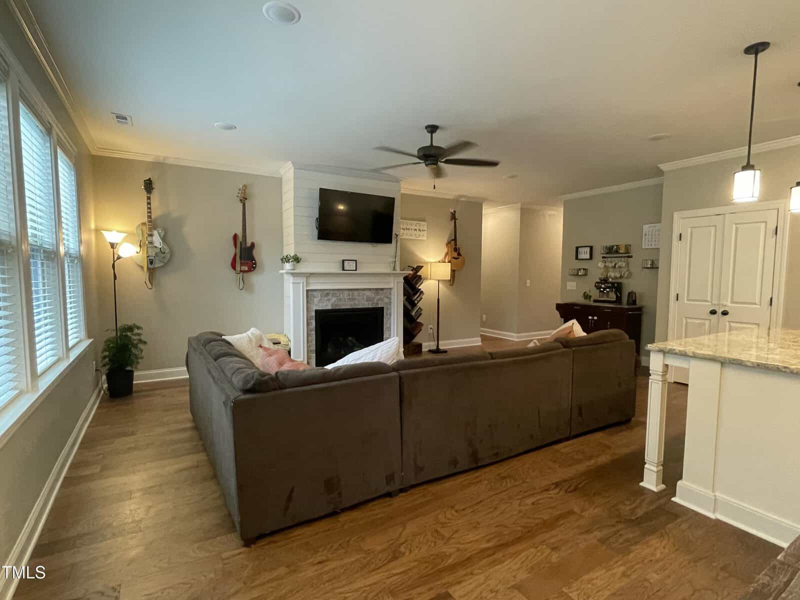 A cozy living room with a brown sectional sofa, wall-mounted TV above a white fireplace, guitars hanging on the walls, hardwood floors, and large windows letting in natural light. A kitchen island is partially visible on the right.