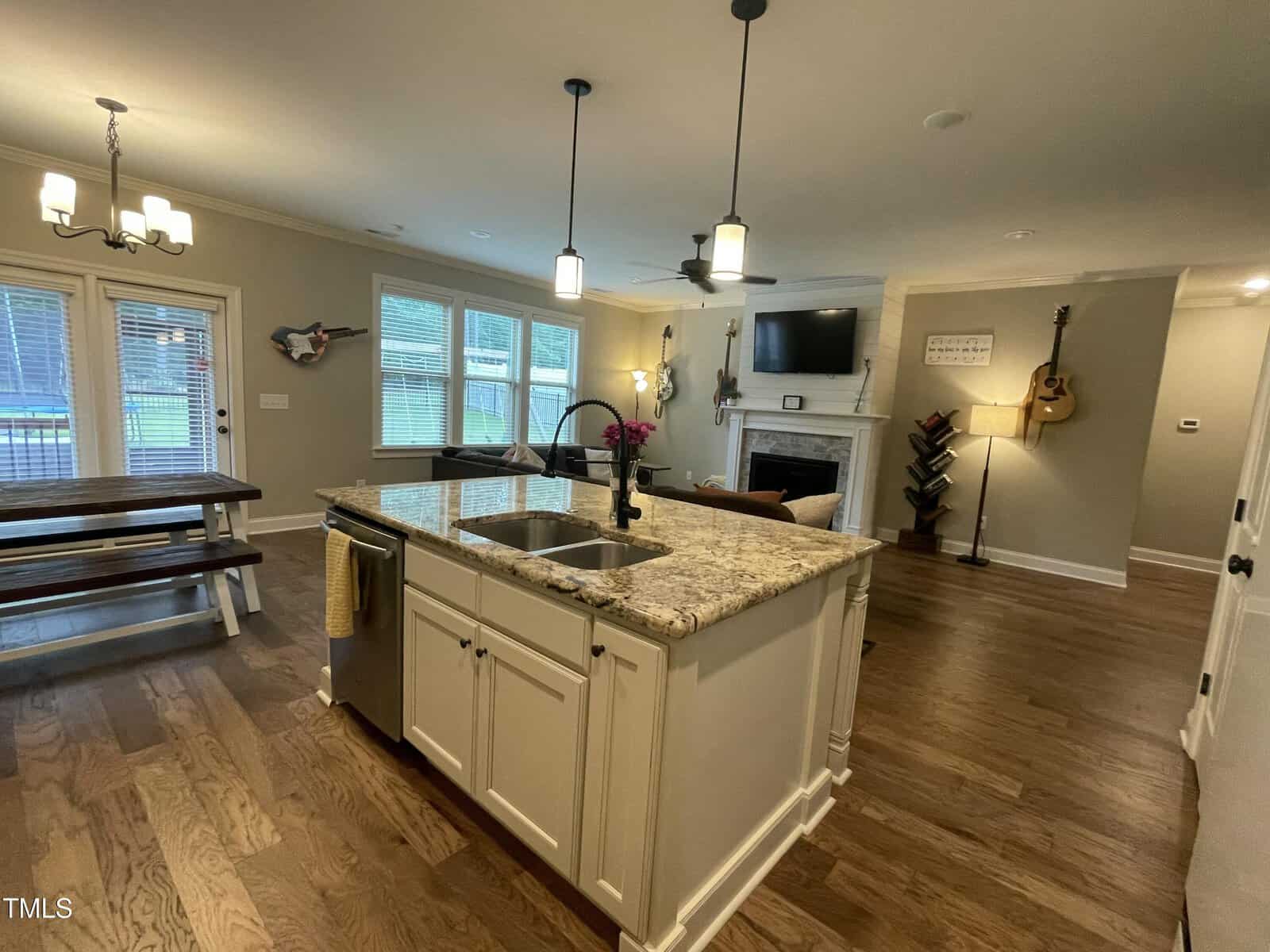 An open kitchen and living area with a center island, dining table, and fireplace. Guitars hang on the walls, and a TV is mounted above the fireplace. Hardwood floors and large windows are visible.