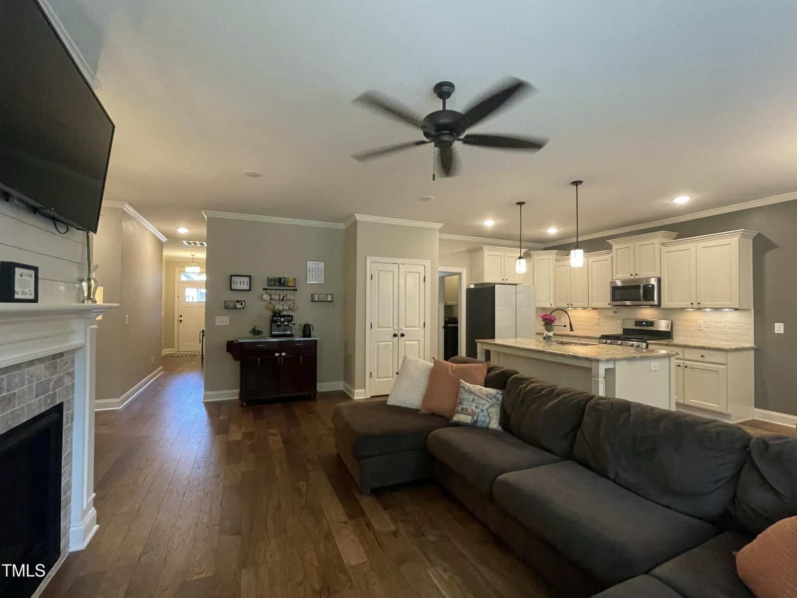Open-concept living area with a dark sectional sofa, ceiling fan, and a kitchen featuring stainless steel appliances, pendant lights, and white cabinetry. Hardwood floors and neutral walls throughout.