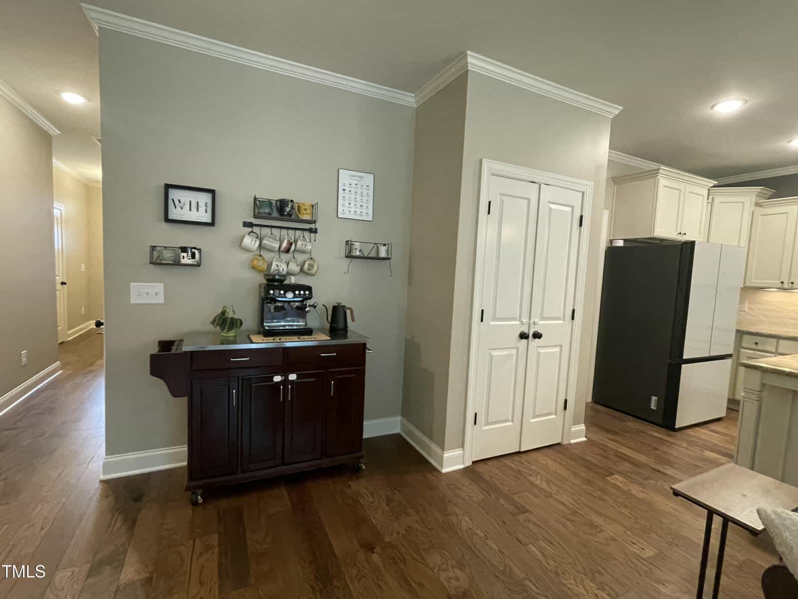 A corner of a kitchen features a dark wood coffee station, wall shelves with mugs, a calendar, and a double white pantry door. Light wood flooring and beige walls are visible, with part of the kitchen and hallway in view.