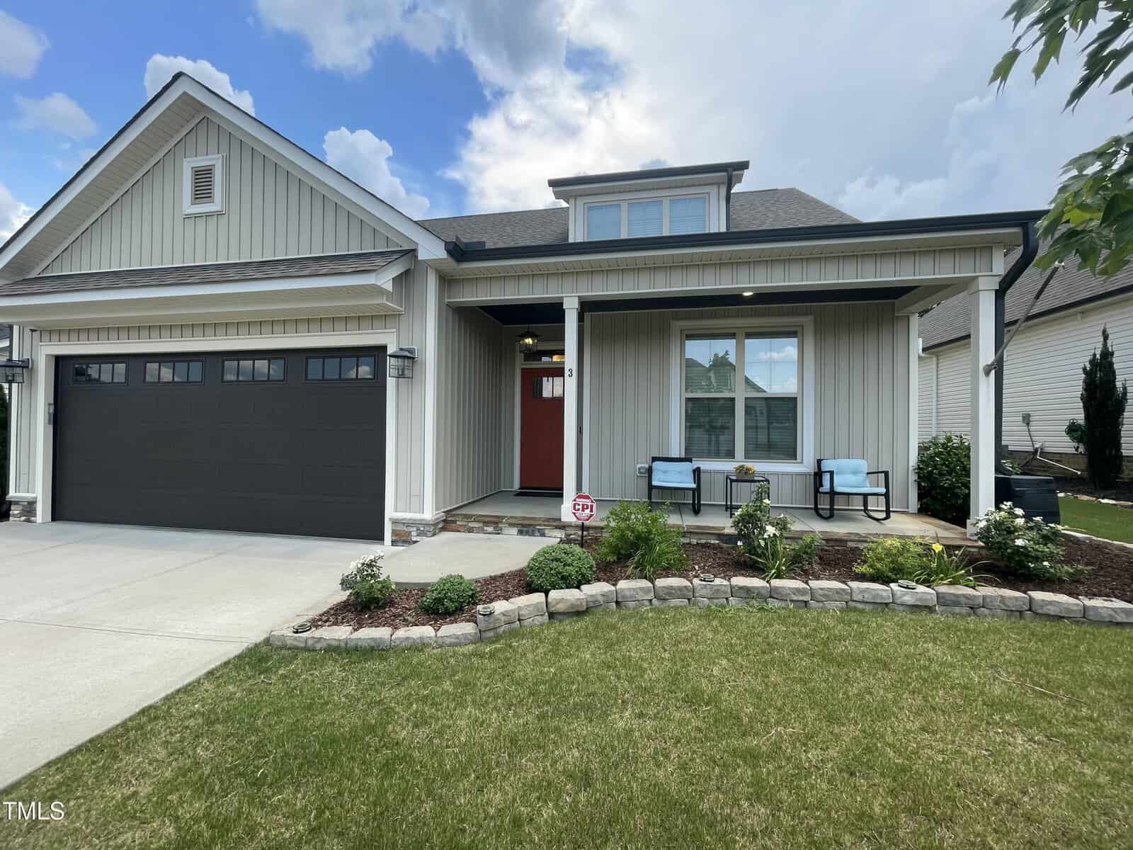 Single-story modern house with a gray garage door, red front door, covered porch, two chairs, and neatly landscaped front yard with shrubs and a stone border under a partly cloudy sky.