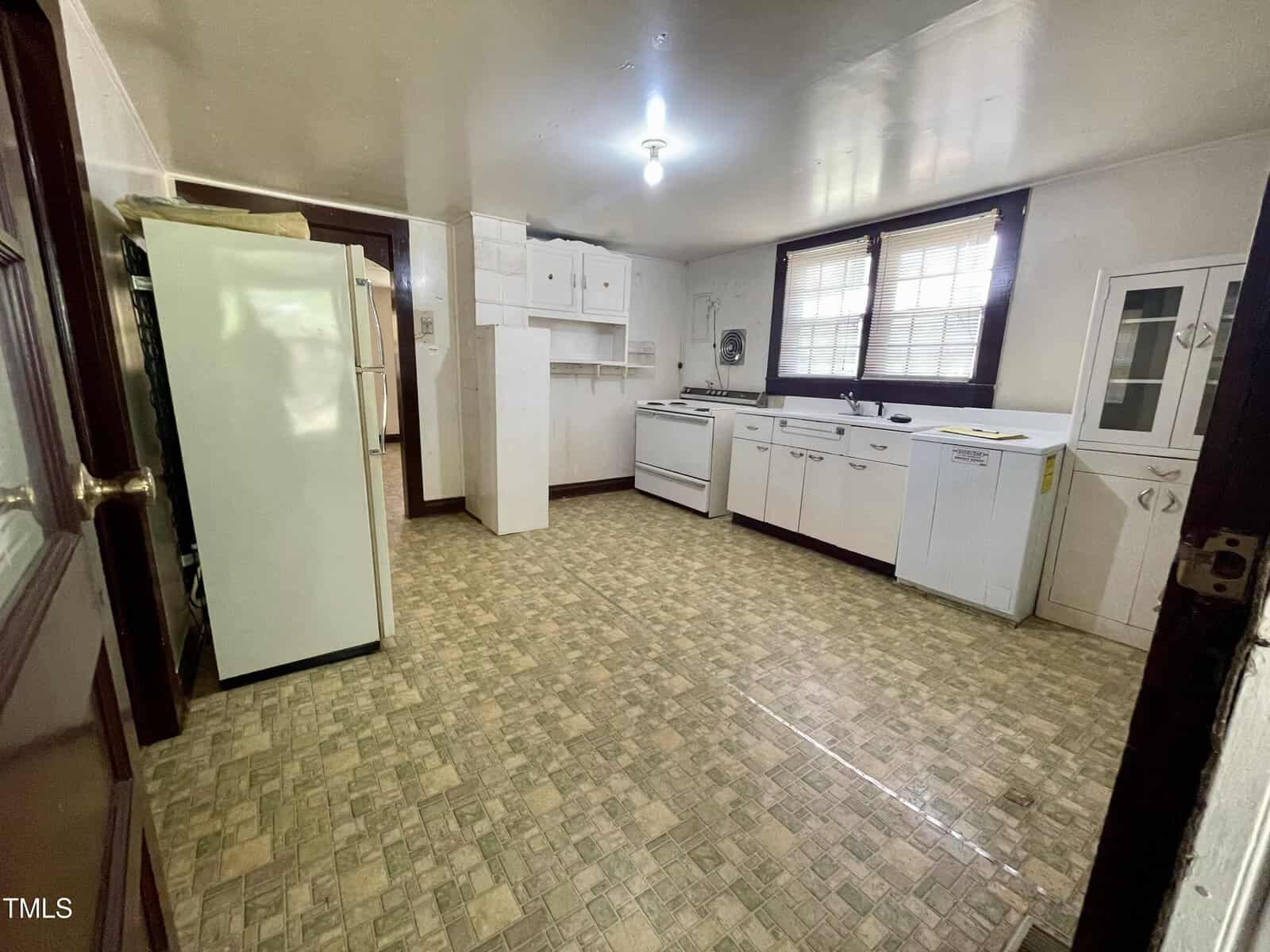 A spacious kitchen with vinyl tile flooring, white cabinets, a refrigerator, a stove, a sink under a window with blinds, and a small wall cabinet. The room is lit by a single ceiling light.