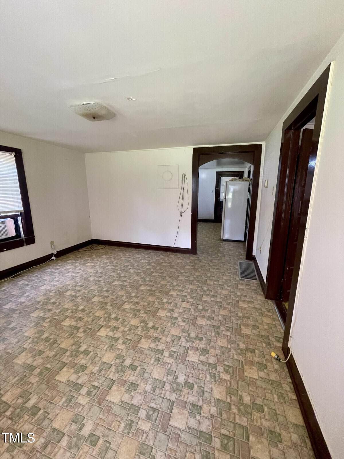 Empty room with tiled floor, cream walls, dark wood trim, and a window on the left. In the background, an open doorway reveals a hallway and a white refrigerator. Ceiling light fixture is missing a cover.