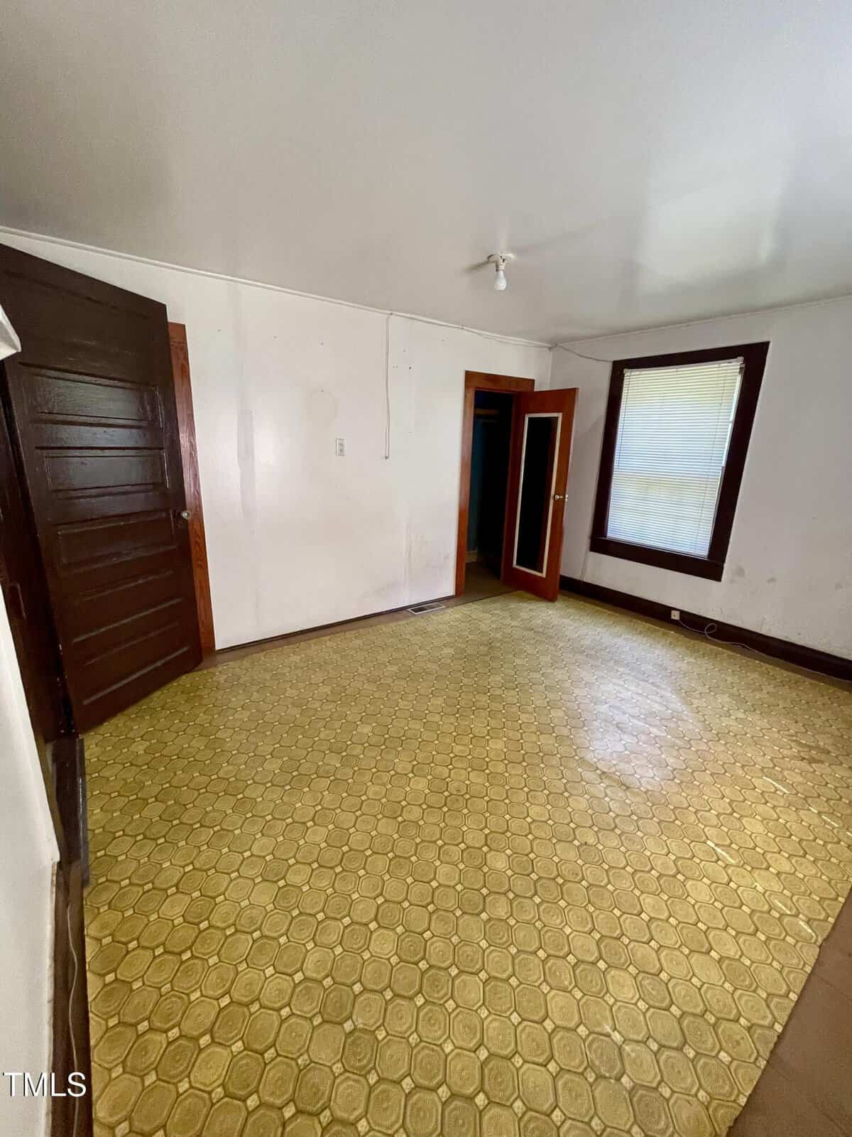 Empty room with yellow patterned linoleum floor, white walls, dark wood trim, a window with blinds, and two open wooden doors. Ceiling light fixture is simple with no shade.