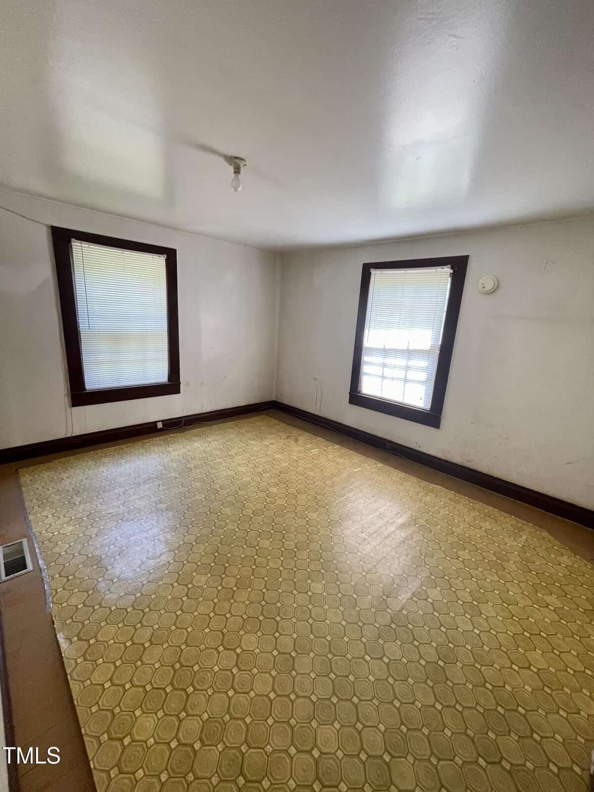 Empty room with two windows, white walls, dark trim, yellow patterned linoleum floor, and a bare ceiling light fixture. Natural light comes through the windows, creating a bright atmosphere.