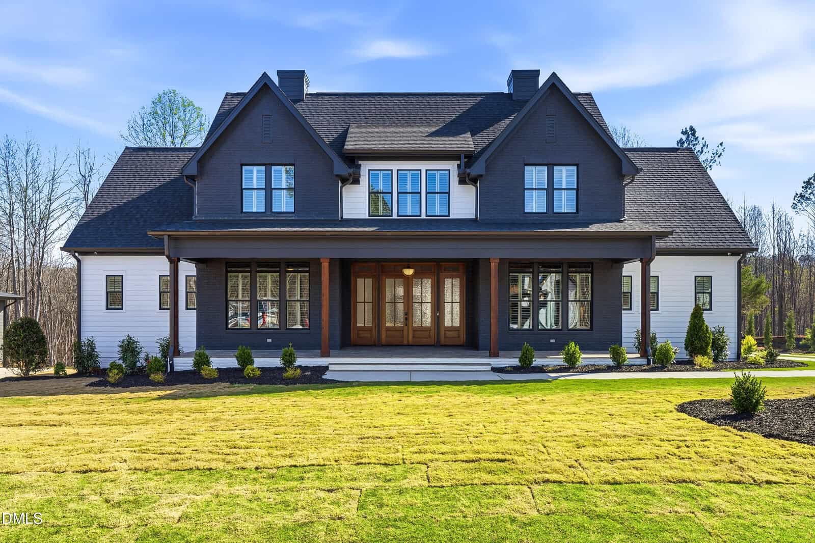 A large, modern two-story house with dark gray and white siding, a covered front porch supported by wooden columns, and large windows. The house is surrounded by a freshly landscaped yard with green grass and small shrubs.
