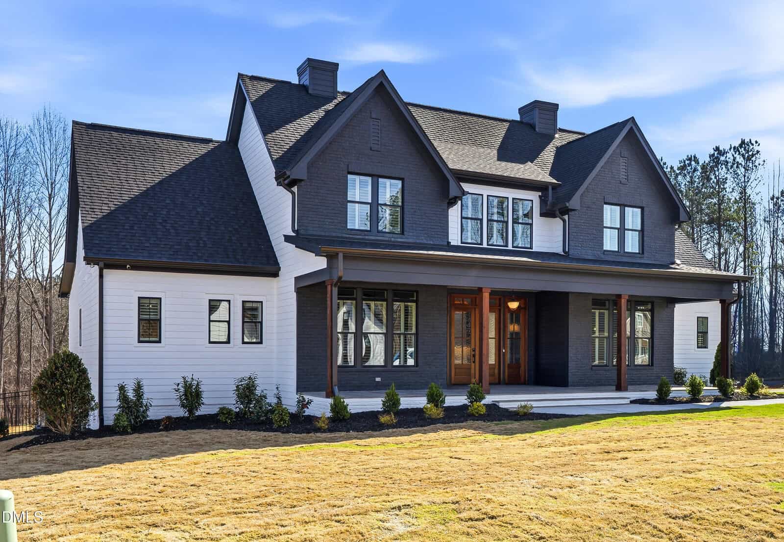 A modern two-story house with dark and white exterior, large front porch with wood columns, black framed windows, and a manicured lawn with shrubs and trees in the background under a blue sky.