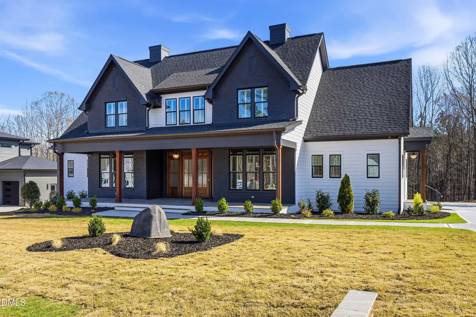 Modern two-story house with dark gray and white exterior, large front porch, double wooden front doors, and a neatly landscaped yard with small bushes and a central boulder. Trees and blue sky in the background.