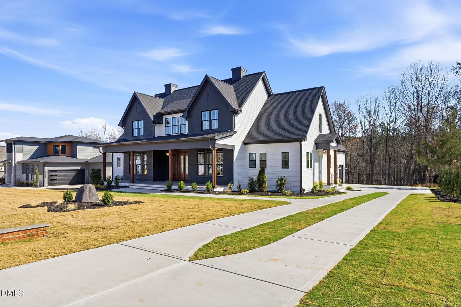 A modern farmhouse-style home with black and white exterior, large front porch, multiple gables, and wide windows. A curved driveway leads to a detached garage, with green lawn and trees in the background.